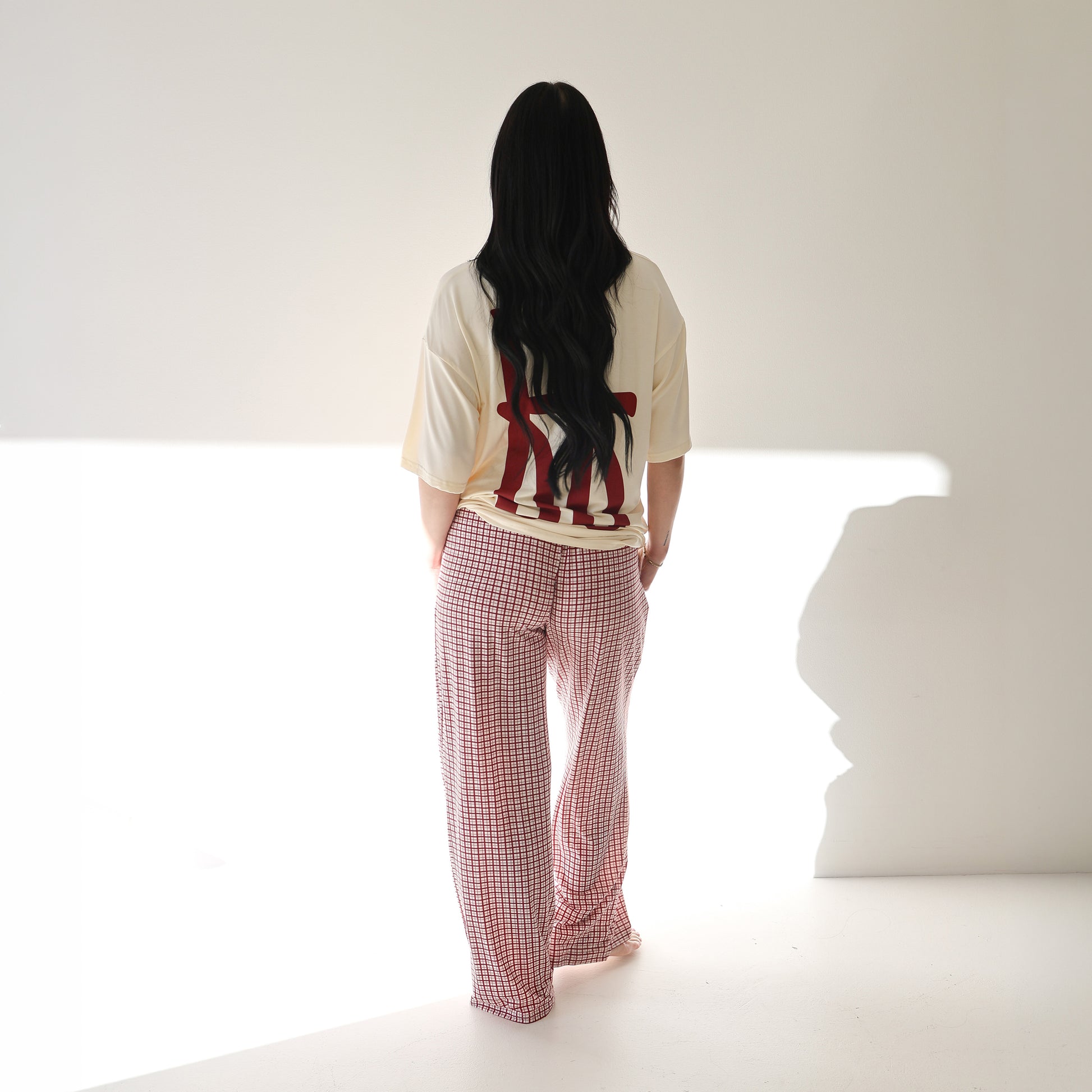 A person with long dark hair stands facing away, wearing the forever french baby Short Sleeve Women's Bamboo Lounge Set in Milk & Window Pane, in a brightly lit, minimal white room.