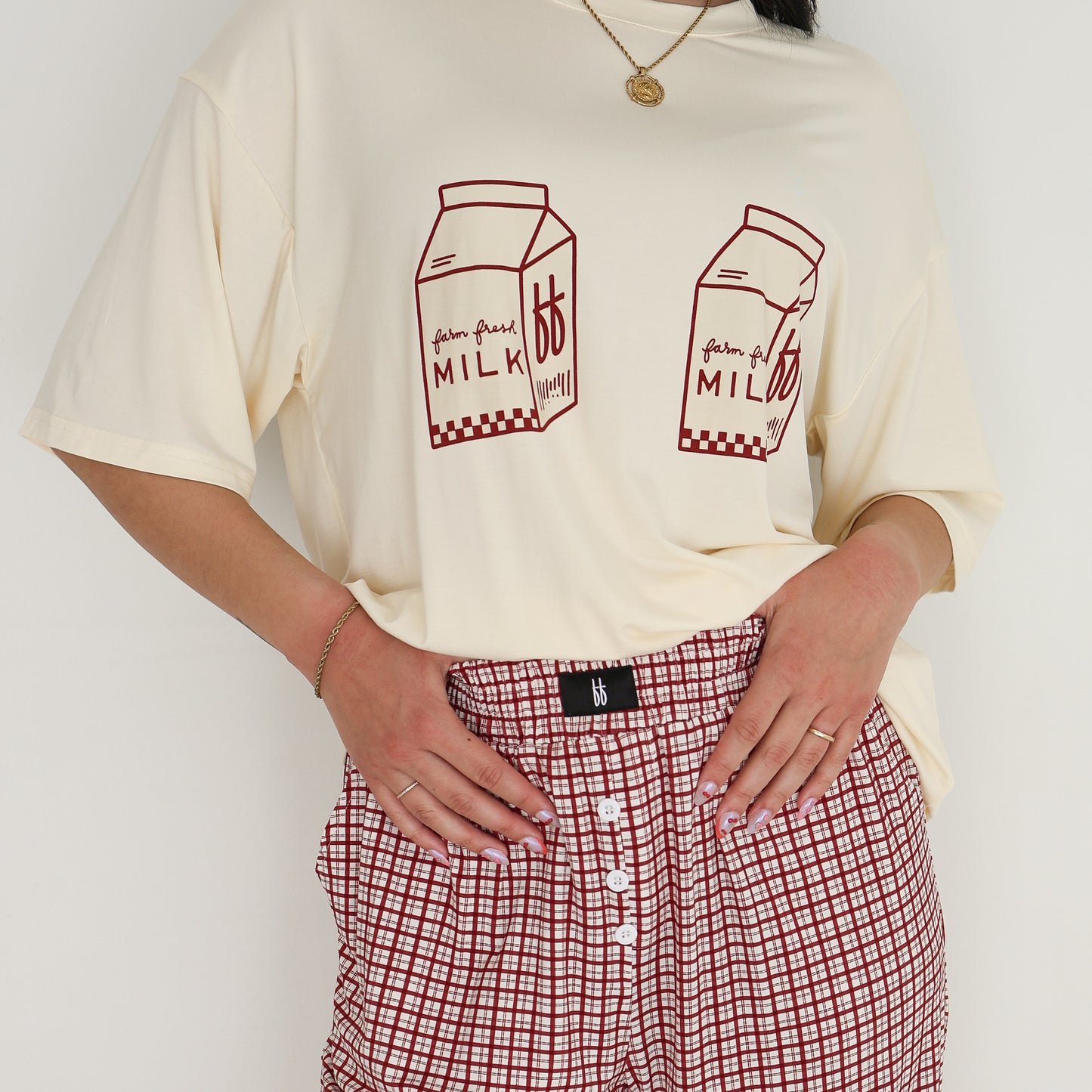 A person wears the forever french baby Short Sleeve Women's Bamboo Lounge Set | Milk & Window Pane, featuring a cream t-shirt with two red milk carton prints and red windowpane-check pants, against a plain white background.