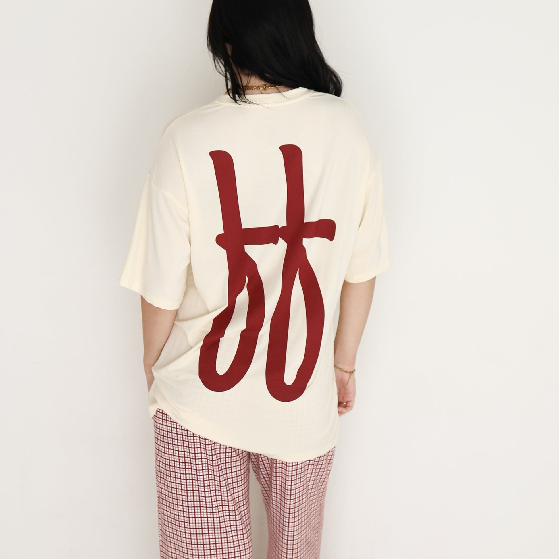 A person with long dark hair stands with their back to the camera, wearing forever french baby's Short Sleeve Women's Bamboo Lounge Set in Milk & Window Pane. The set features a cream top and window pane plaid pants against a plain white background.