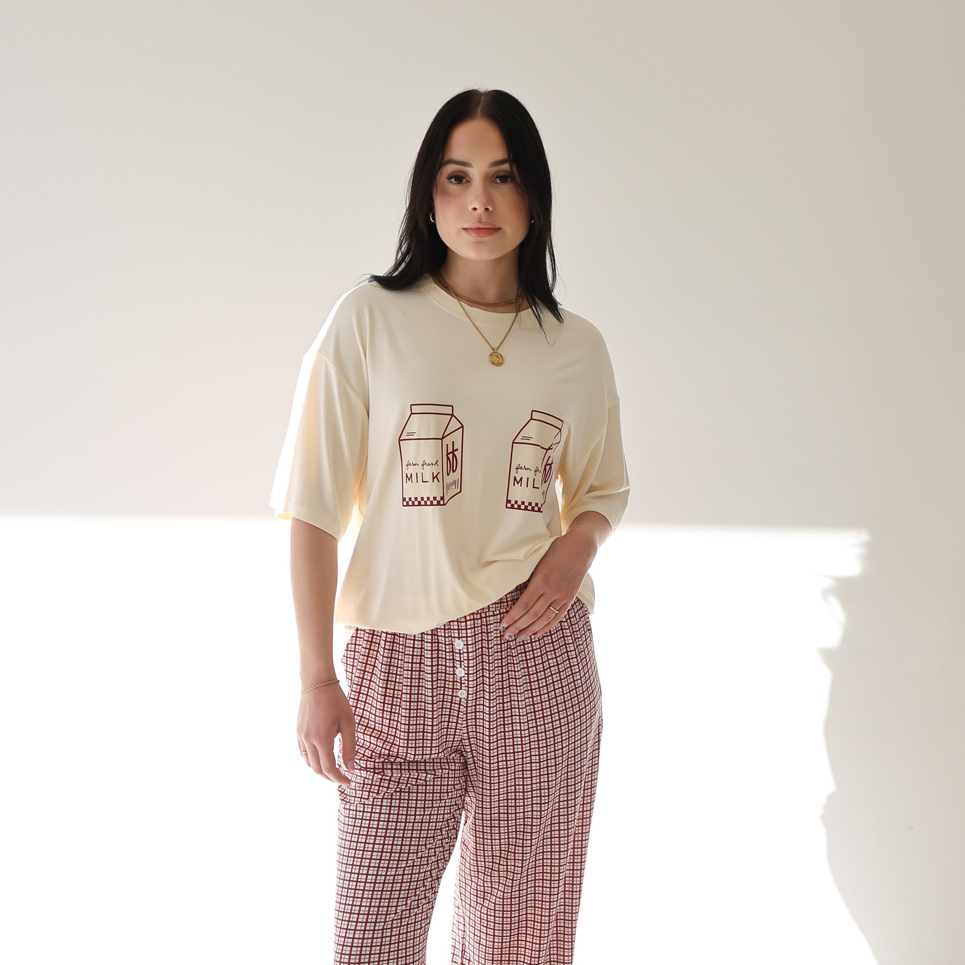 A woman stands against a white wall wearing the forever french baby Short Sleeve Women's Bamboo Lounge Set in Milk & Window Pane, with sunlight casting shadows behind her.