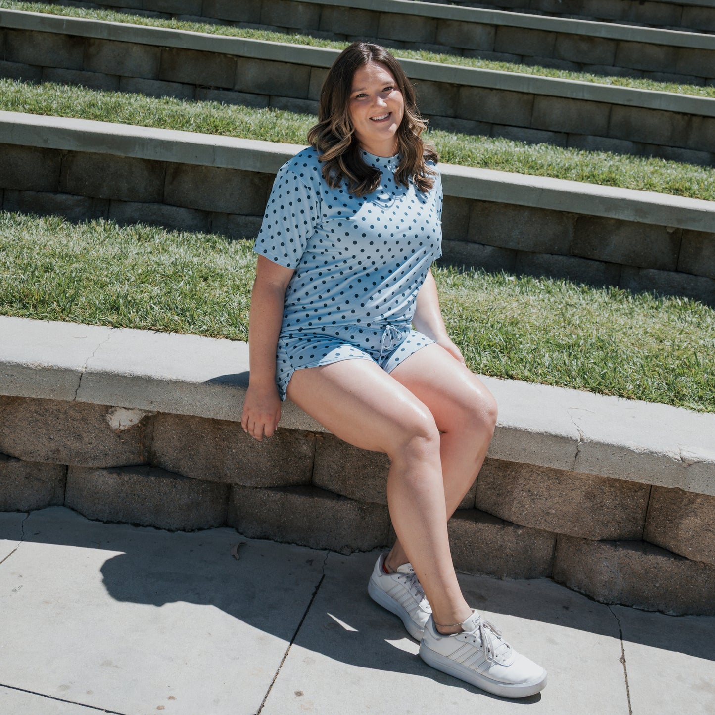 A woman wearing the Women's Bamboo Short Set | Sully by forever french baby sits on a curved stone bench outdoors, smiling. Steps with grass are in the background, and she is wearing white sneakers. This hypo-allergenic bamboo set adds comfort to her relaxed look.