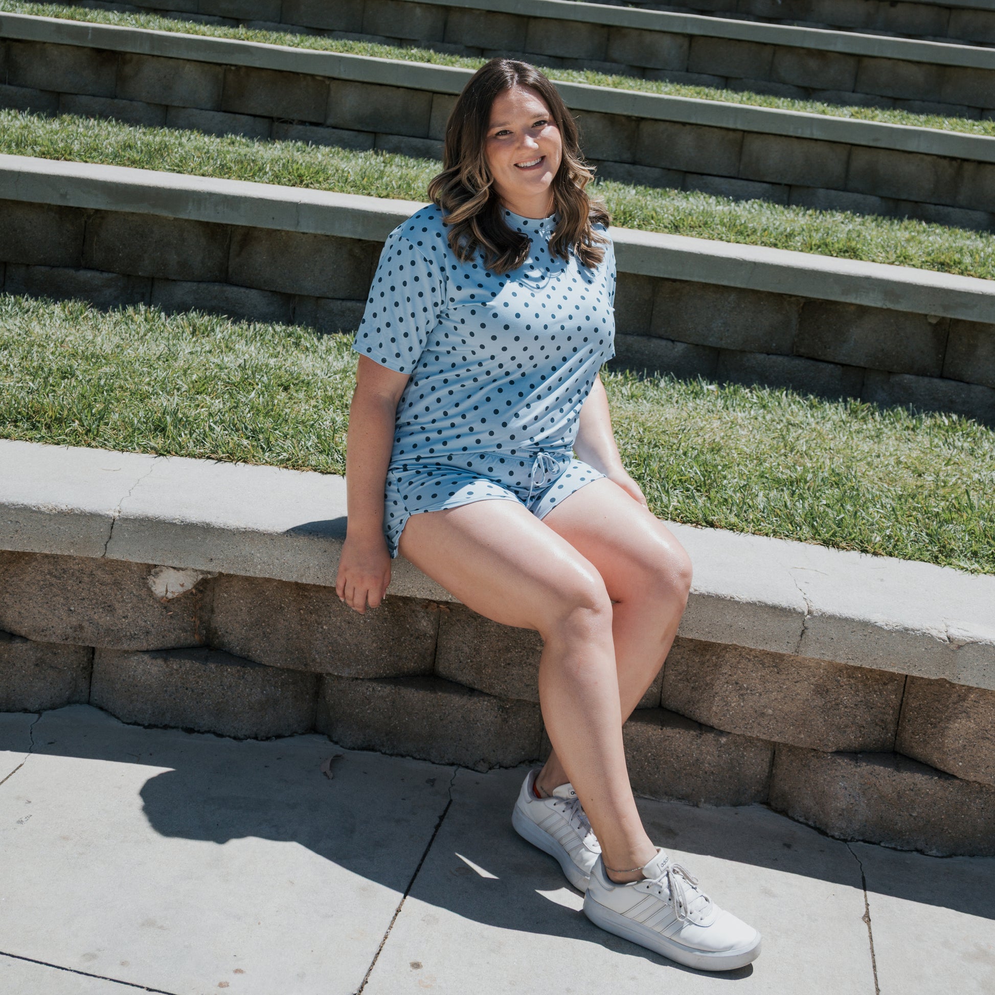 A woman wearing the Women's Bamboo Short Set | Sully by forever french baby sits on a curved stone bench outdoors, smiling. Steps with grass are in the background, and she is wearing white sneakers. This hypo-allergenic bamboo set adds comfort to her relaxed look.