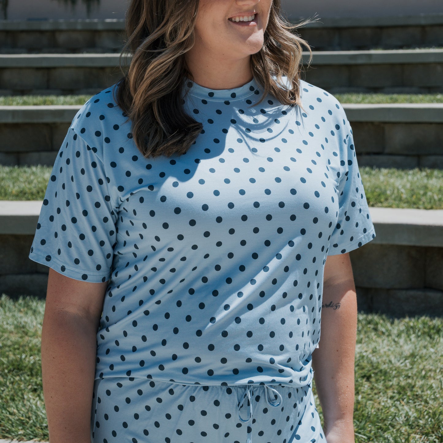 A person wearing the forever french baby Women's Bamboo Short Set | Sully—a light blue, hypo-allergenic pajama set with black polka dots—stands outside on a sunny day, with stone steps and grass in the background.