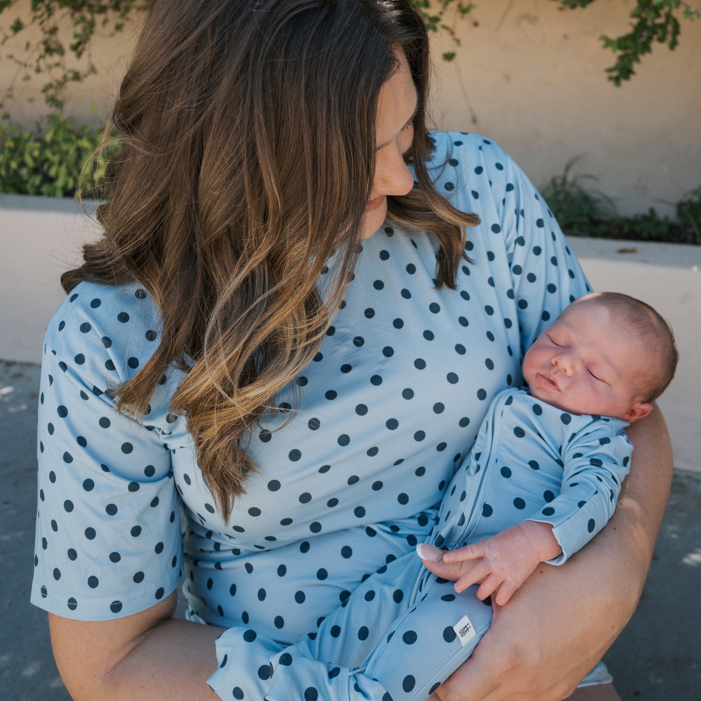 A woman with wavy brown hair cradles a sleeping baby in her arms, both dressed in matching forever french baby Women’s Bamboo Short Set | Sully pajamas—a light blue, black polka-dotted set crafted from soft, hypo-allergenic bamboo for optimal comfort. They stand outside near a light-colored wall adorned with green plants.