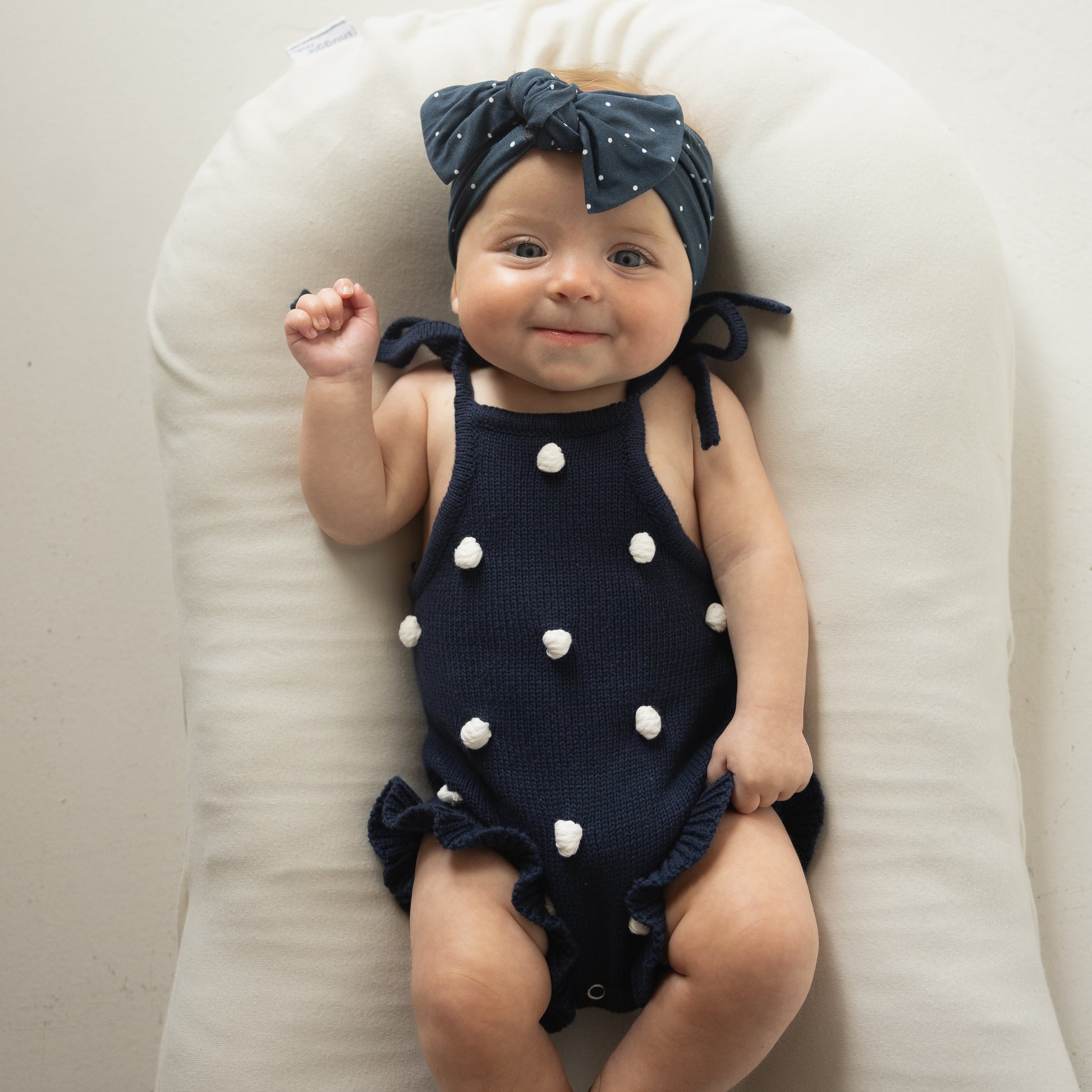 A baby wearing the forever french baby Knit Ruffle Romper | Emery with adjustable tie straps, white pom-poms, and a matching navy headband with white dots, lies smiling on a white cushioned surface.