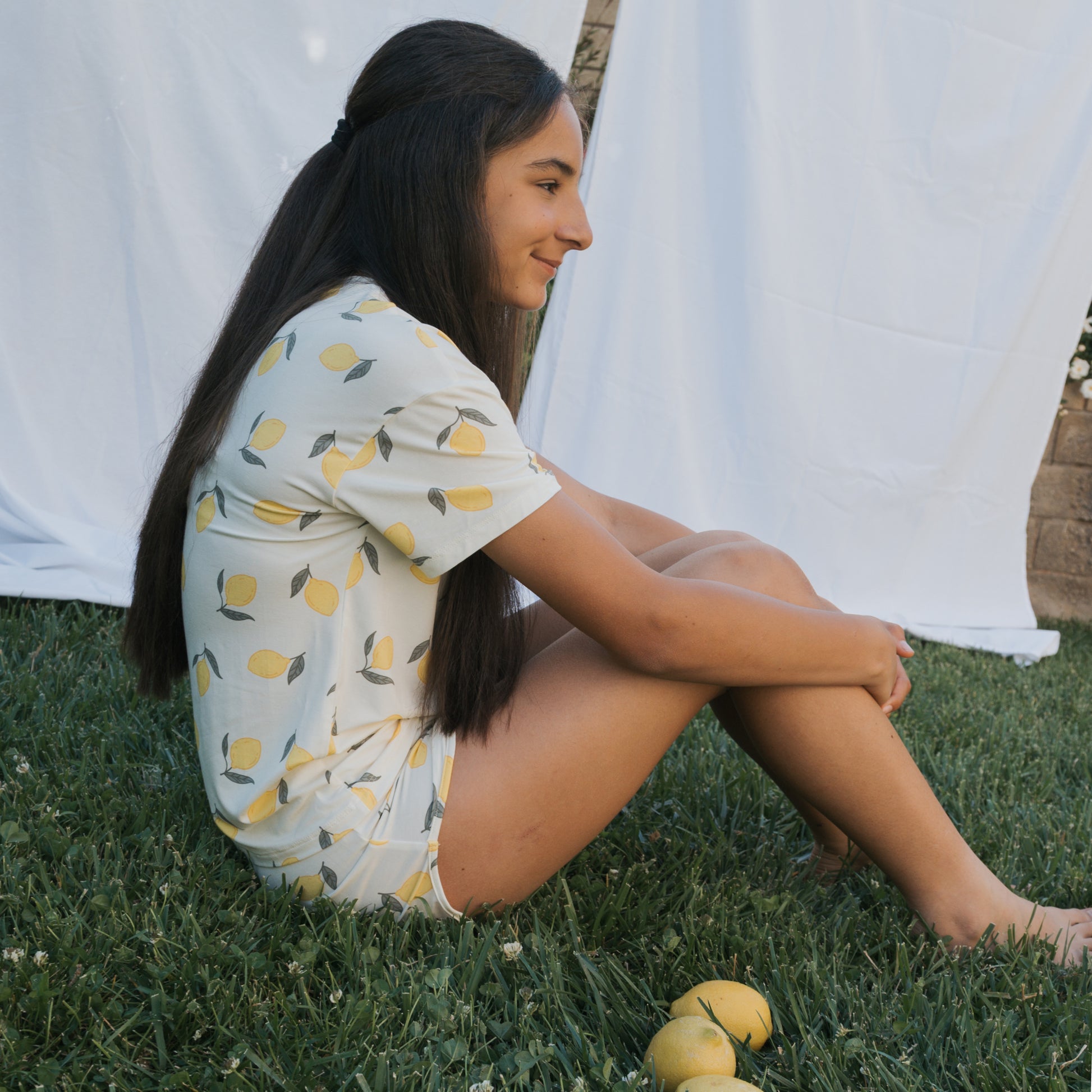 A person with long dark hair sits cross-legged on grass, wearing the forever french baby Girl's Bamboo Short Set | Lemon. Hypo-allergenic material adds comfort, with lemons nearby and white fabric in the background.