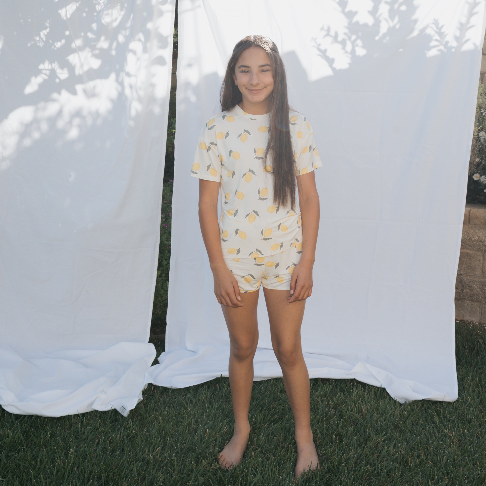 A young girl stands barefoot on grass before white sheets outside, smiling in the forever french baby Girl's Bamboo Short Set | Lemon. Sunlight highlights her hypo-allergenic, comfy bamboo outfit featuring a cheerful lemon print.