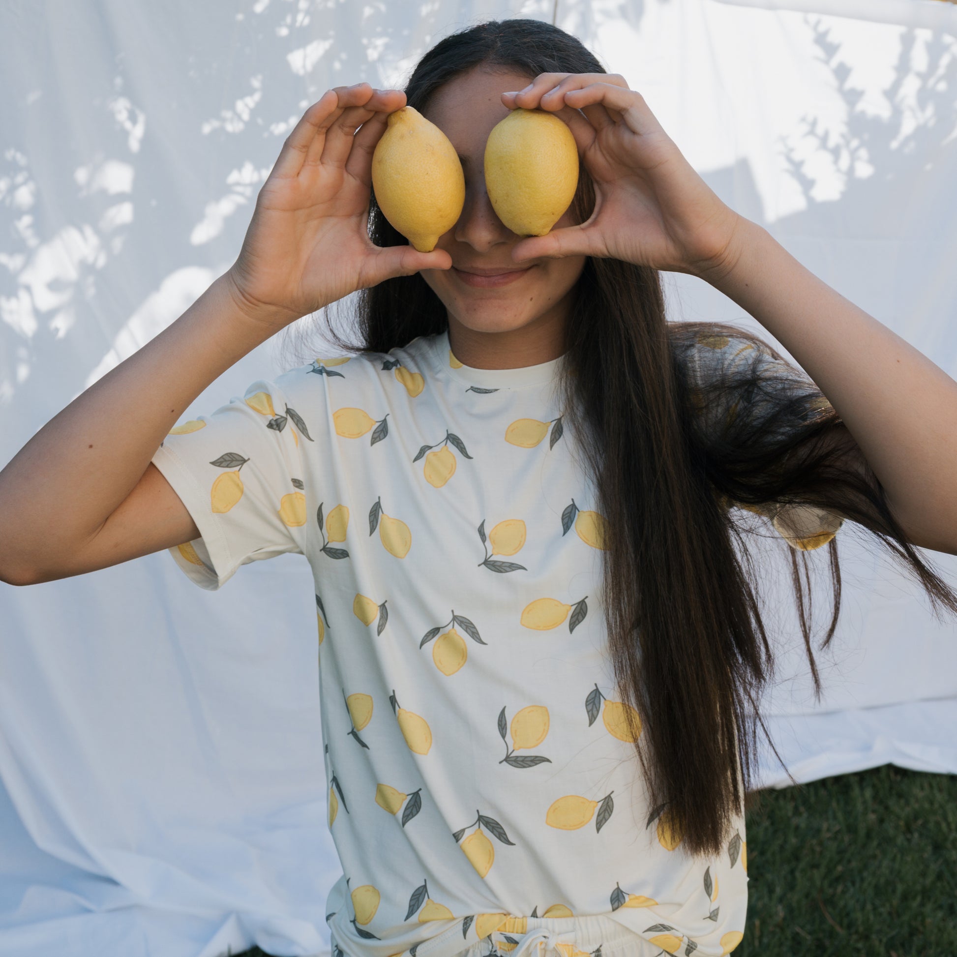 A smiling girl with long dark hair holds two lemons over her eyes, wearing the forever french baby Girl's Bamboo Short Set | Lemon. She stands outdoors in front of a sun-dappled white sheet backdrop with tree shadows.