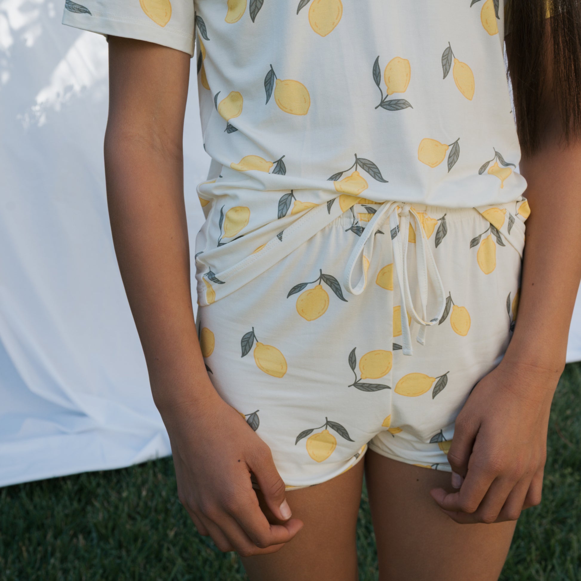 A girl models the forever french baby Girl's Bamboo Short Set | Lemon, featuring a yellow lemon print, standing on grass with hands by her sides. A white fabric backdrop is behind her.
