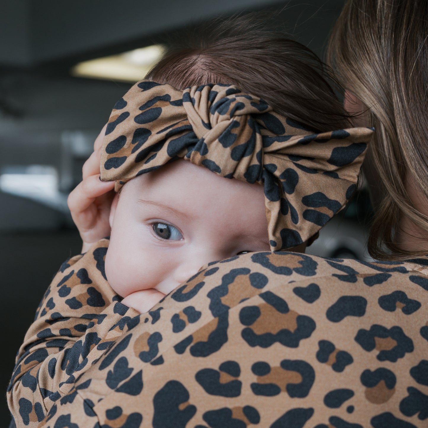 A baby wearing the forever french baby Bamboo Head Wrap | Leopard rests on a woman’s shoulder, both dressed in matching leopard print. The baby looks over her shoulder, gently sucking on their hand.