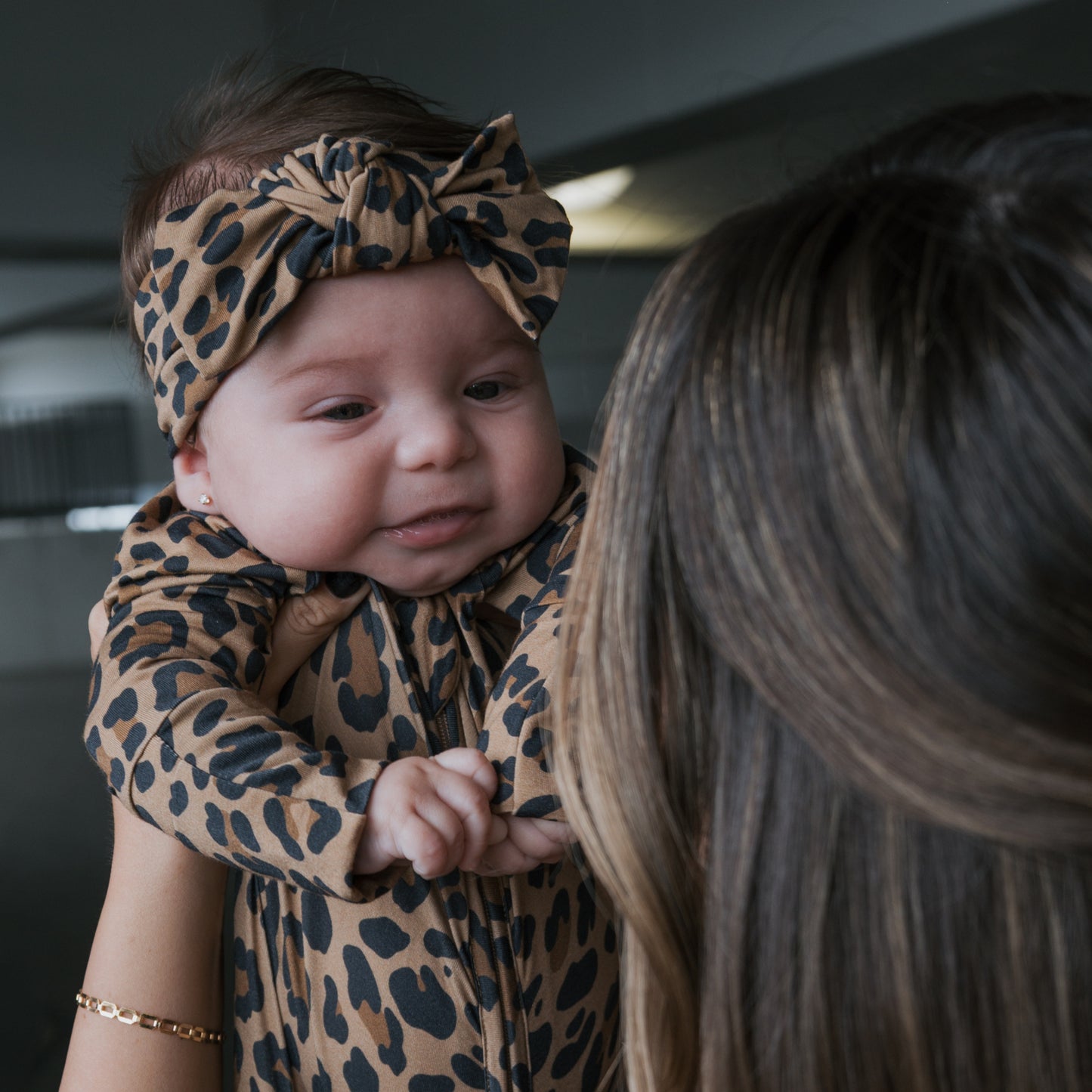 A baby wearing the forever french baby Bamboo Head Wrap | Leopard is held by an adult with long brown hair. The baby's gentle smile and the softly blurred background highlight the adorable head wrap.