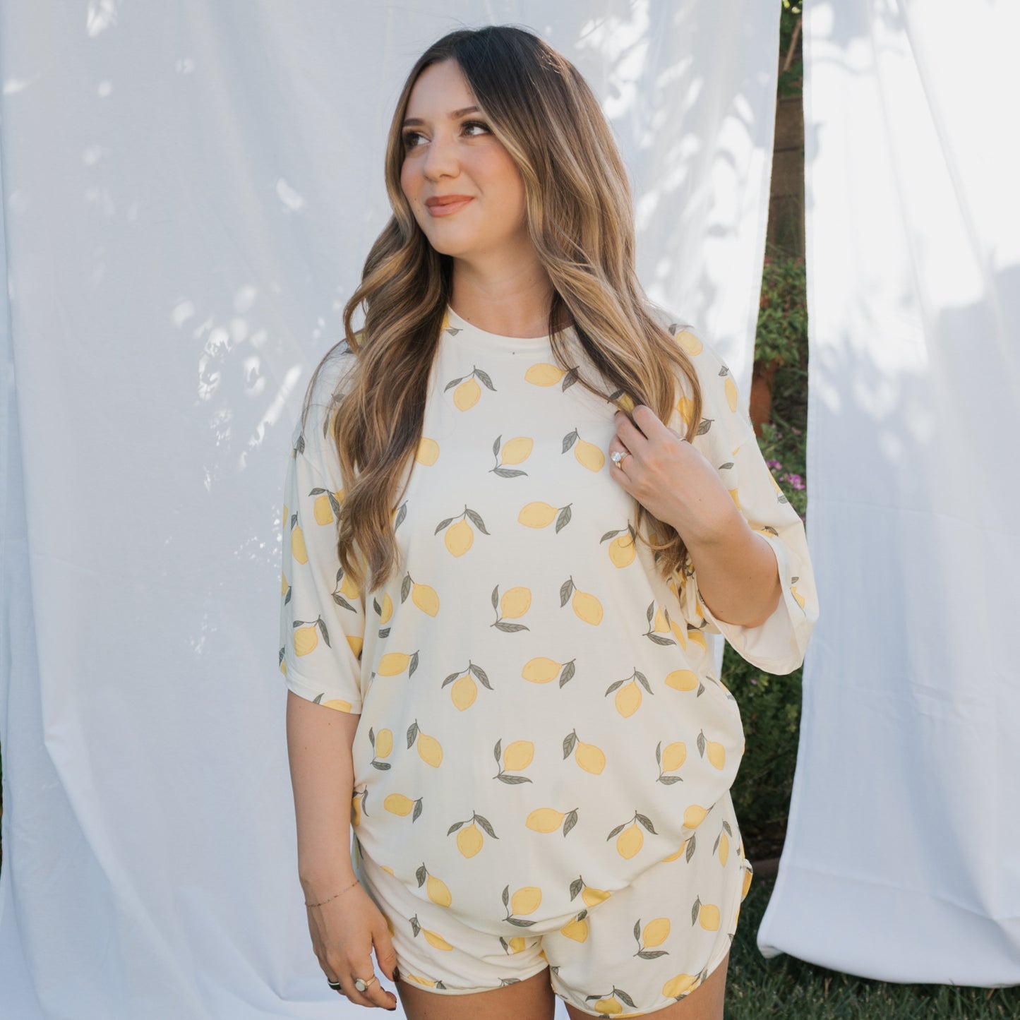 A woman with long, wavy hair smiles softly outdoors in front of white fabric backdrops, wearing the forever french baby Women's Bamboo Short Set | Lemon.