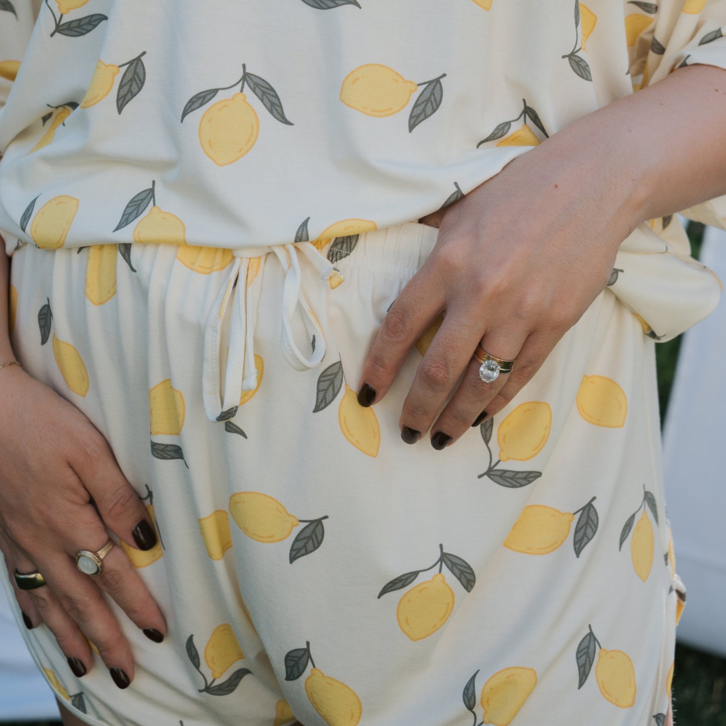 A person wears the Women's Bamboo Short Set | Lemon by forever french baby—an oversized, cream-colored bamboo t-shirt and shorts set with a lemon print—standing hands on waist, showing several rings and dark nail polish.