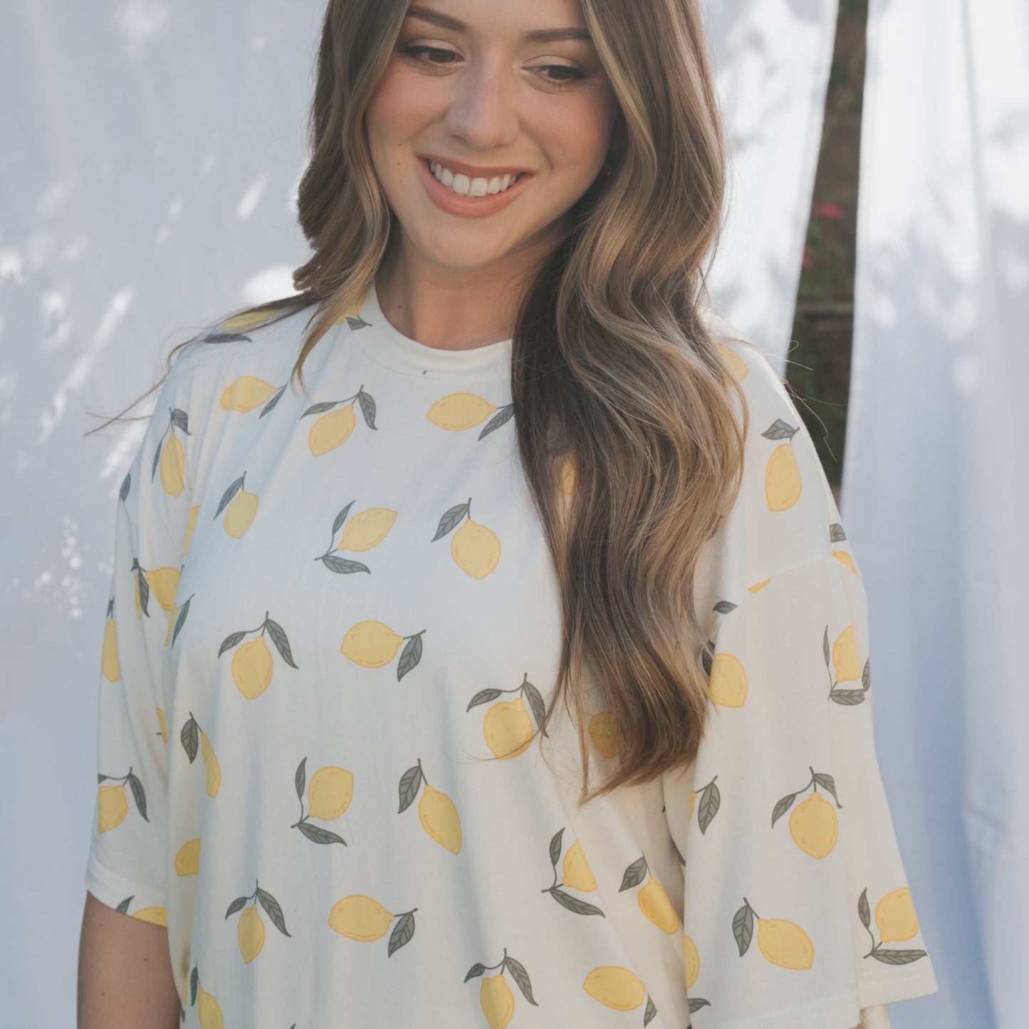 A woman with long, wavy brown hair smiles while wearing the forever french baby Women's Bamboo Short Set | Lemon, a hypo-allergenic white outfit with a yellow lemon pattern. White curtains and soft natural light appear in the background.