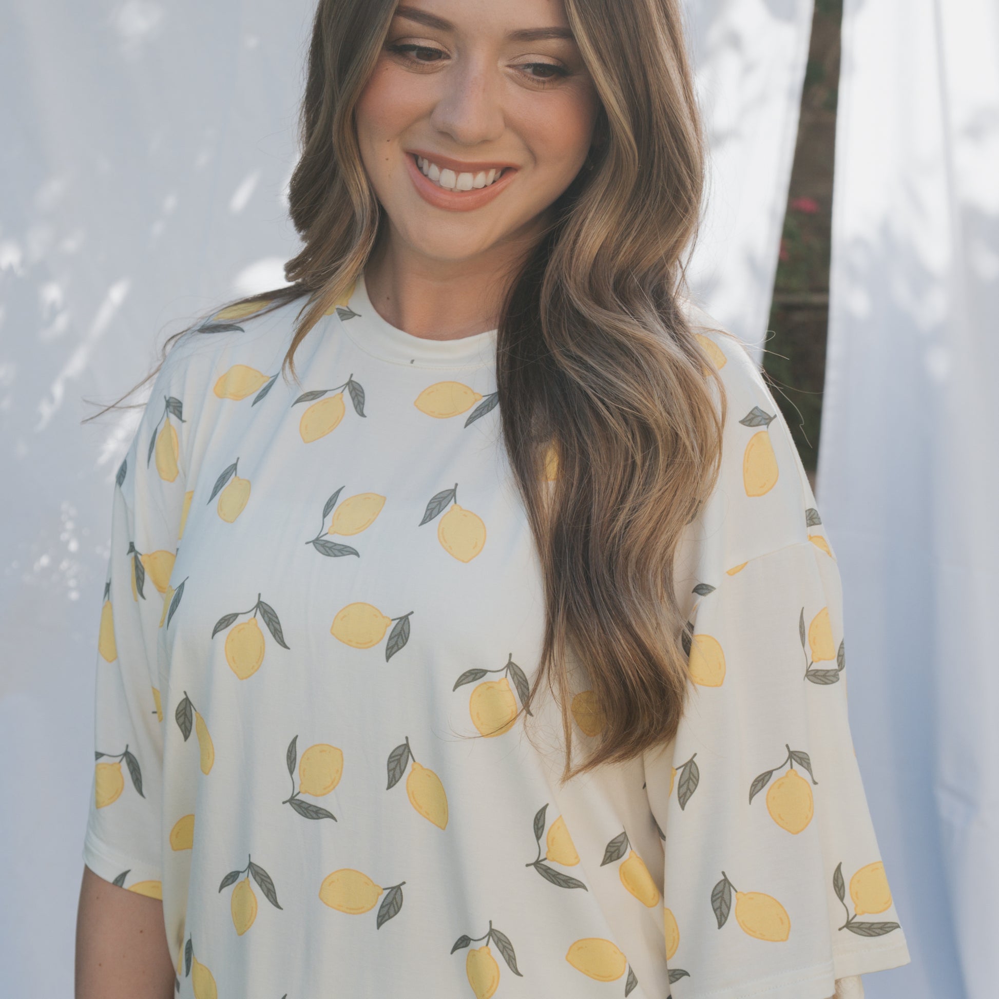 A woman with long, wavy brown hair smiles while wearing the forever french baby Women's Bamboo Short Set | Lemon, a hypo-allergenic white outfit with a yellow lemon pattern. White curtains and soft natural light appear in the background.