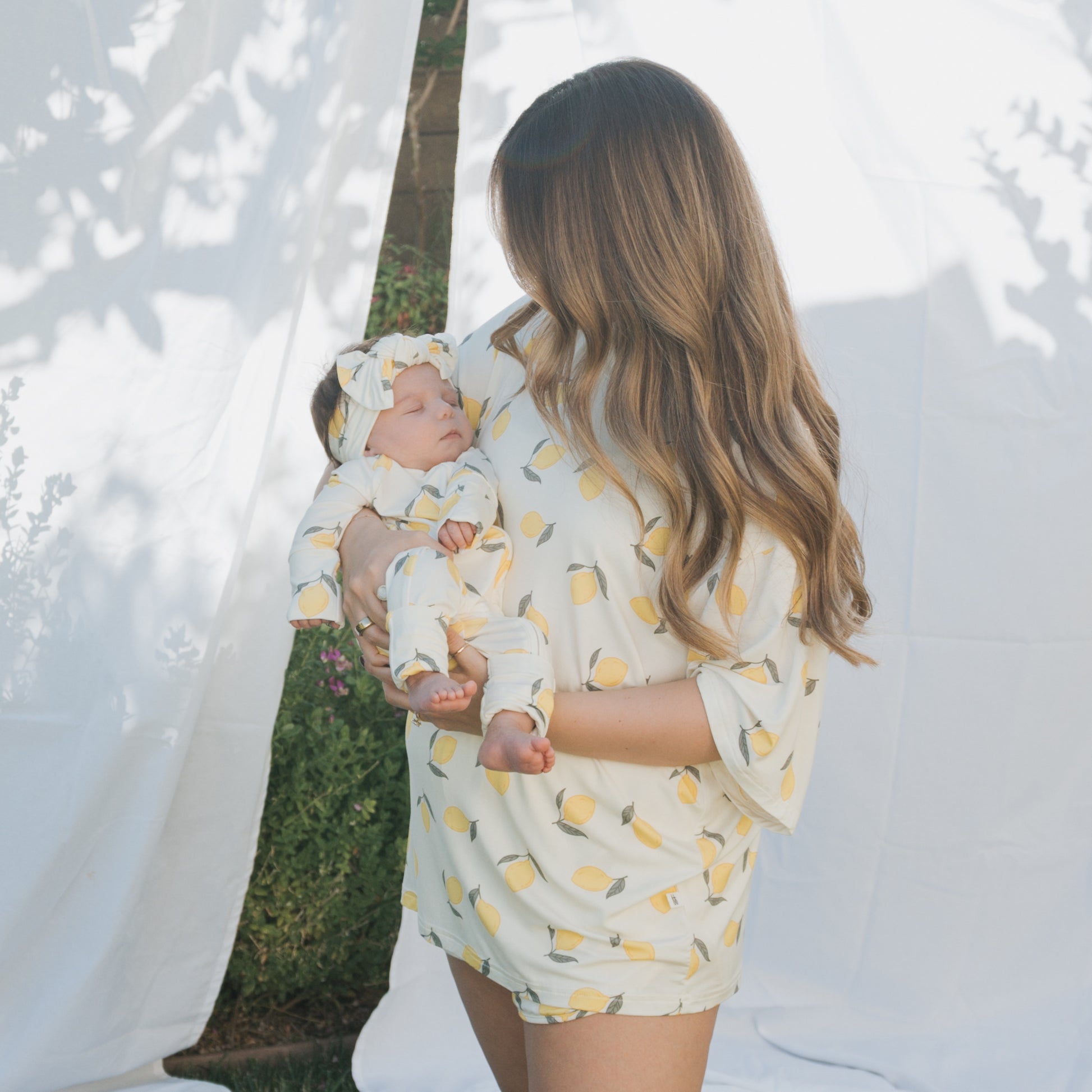 A woman with long brown hair holds a baby outdoors, both wearing forever french baby's Women's Bamboo Short Set | Lemon—matching cream bamboo outfits with yellow lemon prints—beside white fabric panels in sun and shadow.