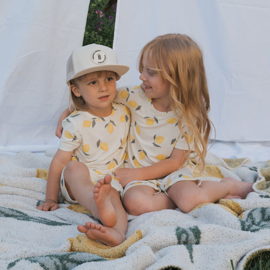 Two young children sit outside on a blanket in matching Bamboo Short Sets | Lemon from forever french baby; one wears a white hat, looking ahead, while the other smiles with an arm around them. White sheets hang in the background.