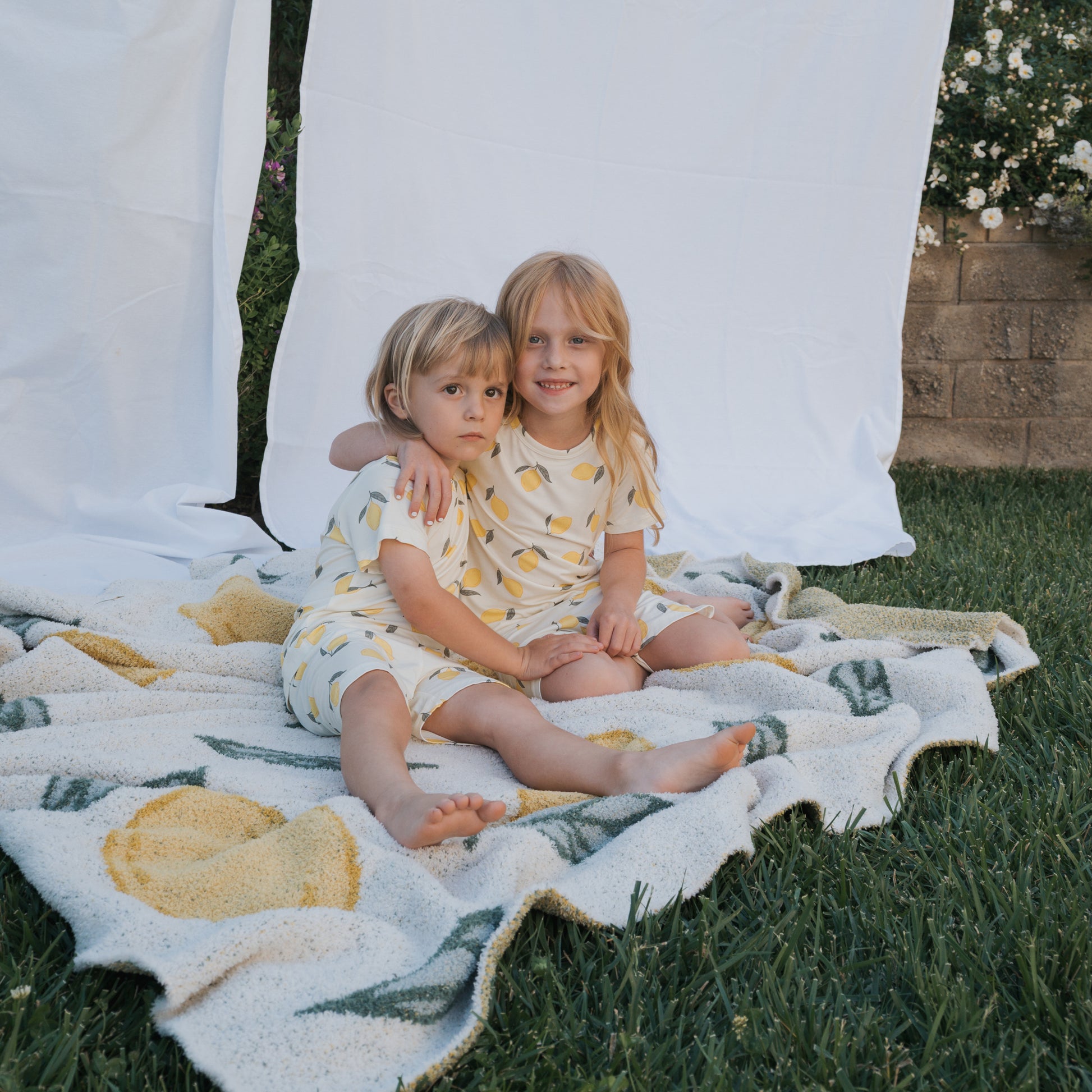 Two young children sit on a lemon-patterned blanket, wearing matching forever french baby Bamboo Short Sets in Lemon. One smiles and hugs the other, who looks serious. White sheets hang behind them and flowers bloom along a brick wall.