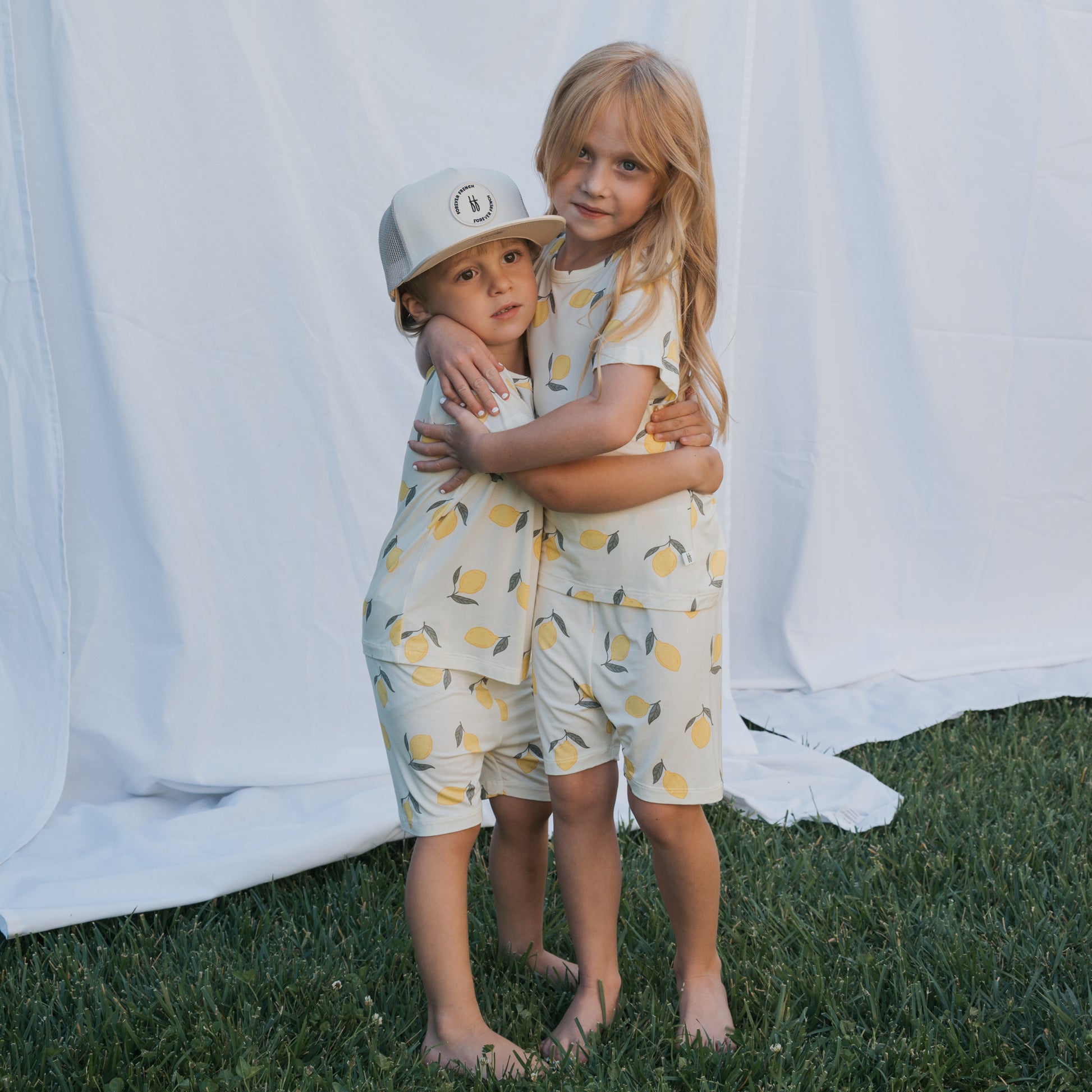 Two young children wearing forever french baby Bamboo Short Set | Lemon pajamas stand barefoot on grass and hug each other. One wears a white cap. A white sheet in the background highlights their breathable sleepwear.