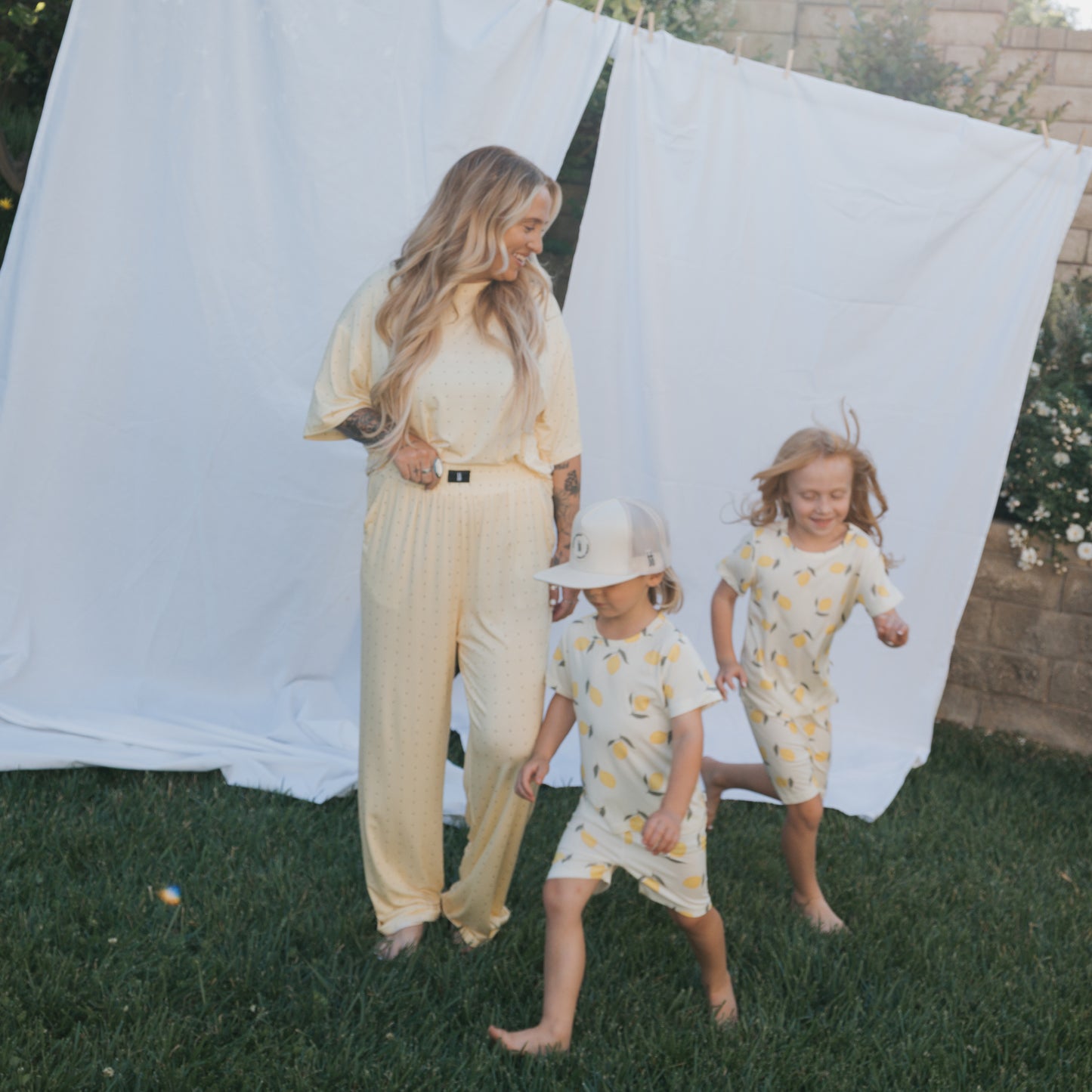A woman in light yellow stands on grass before a white sheet, smiling at two young children in matching forever french baby Bamboo Short Set | Lemon, who play and run barefoot in their hypoallergenic kids’ clothing.