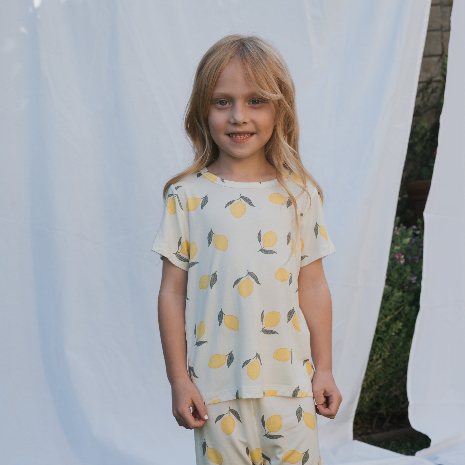 A young girl with long blonde hair smiles in front of a white sheet backdrop, wearing the forever french baby Bamboo Short Set | Lemon. The breathable pajamas feature a yellow lemon print, with green plants visible in the background.