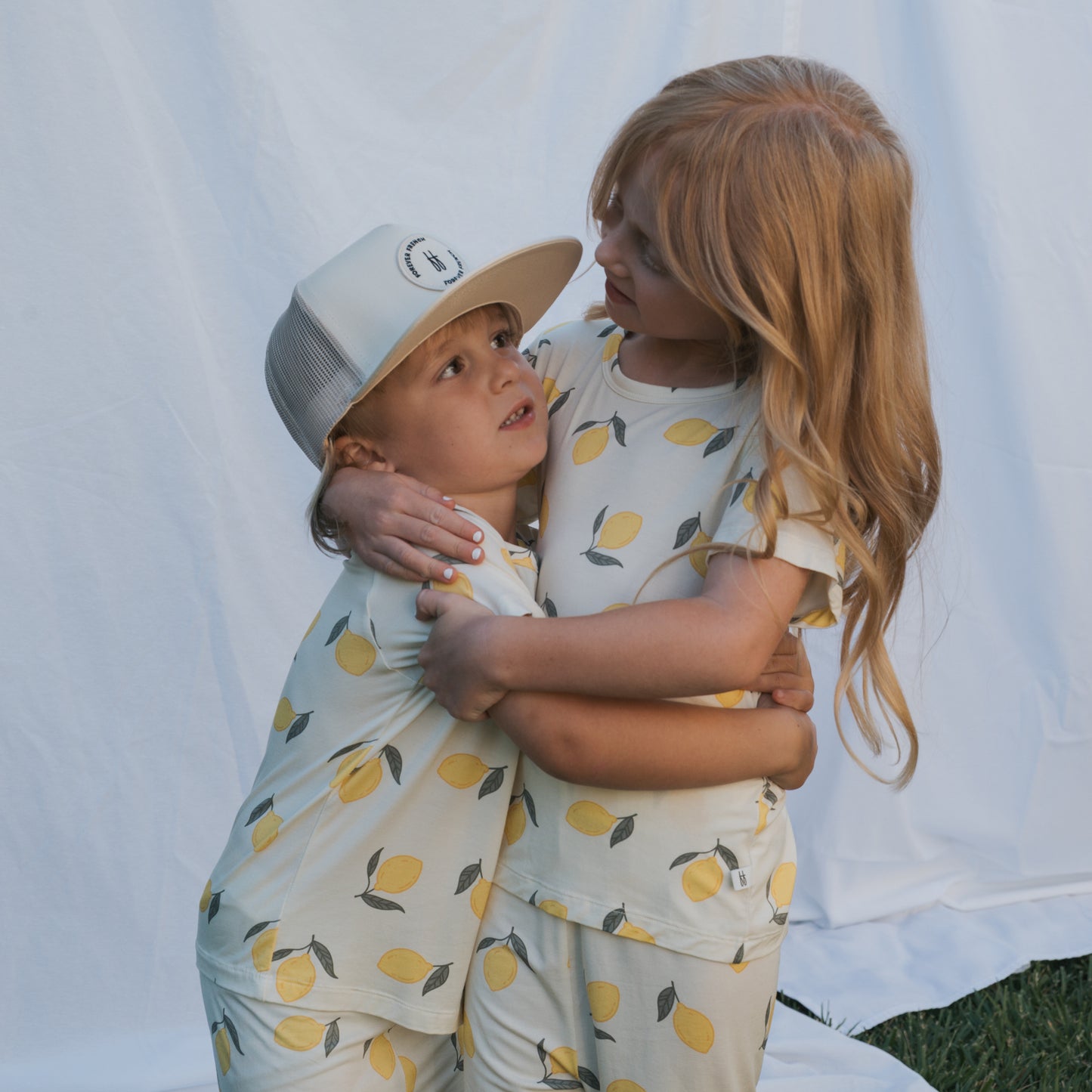 Two young children in matching Bamboo Short Set | Lemon pajamas by forever french baby hug in front of a white backdrop—one wears a cap, the other smiles—showcasing comfy, hypoallergenic kids’ clothing perfect for any moment.
