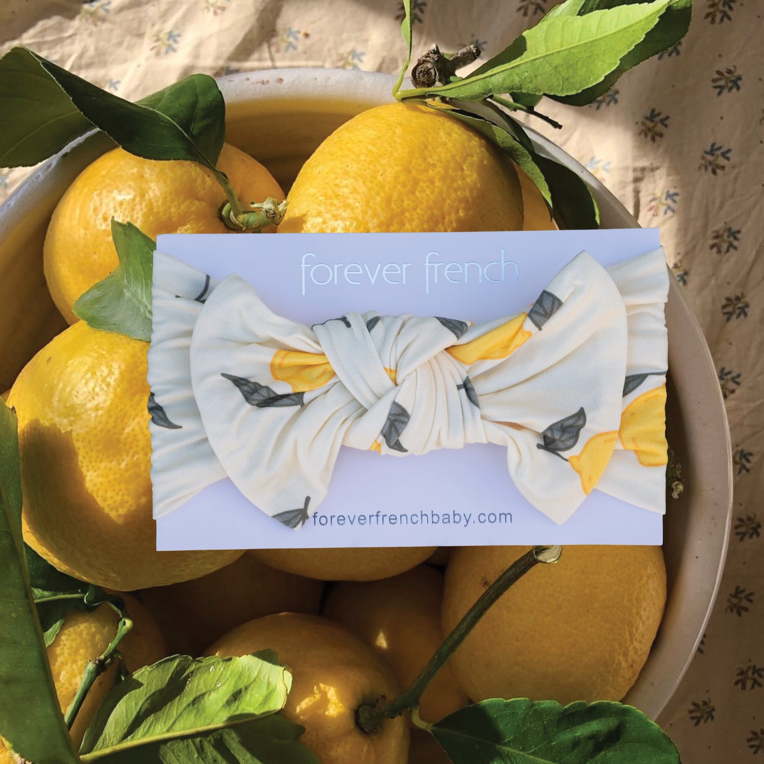 A bowl of lemons with green leaves rests on a patterned cloth. Featured is the forever french baby Bamboo Head Wrap | Lemon, showcasing its lemon print and the website foreverfrenchbaby.com.