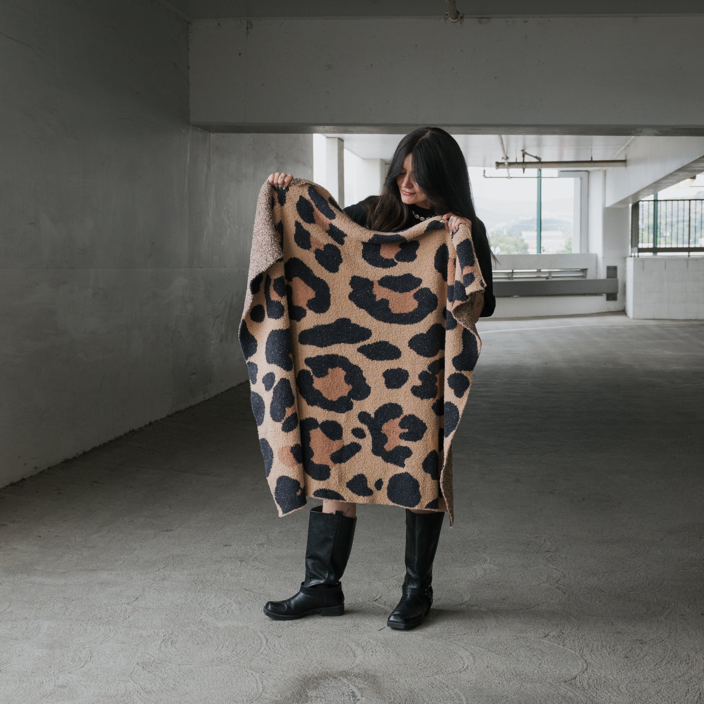 In a concrete parking garage, a person with long dark hair and black boots holds the forever french baby Plush Blanket | Leopard—an OEKO-TEX certified beige and black leopard-print soft blanket—partially wrapped around themselves.