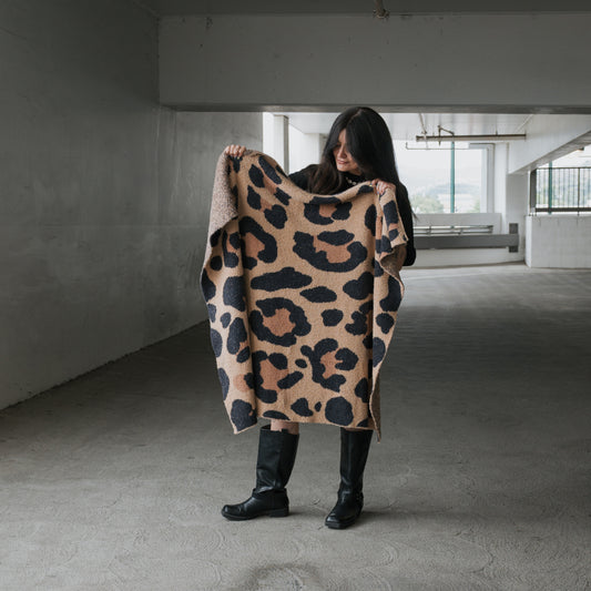 In a concrete parking garage, a person with long dark hair and black boots holds the forever french baby Plush Blanket | Leopard—an OEKO-TEX certified beige and black leopard-print soft blanket—partially wrapped around themselves.