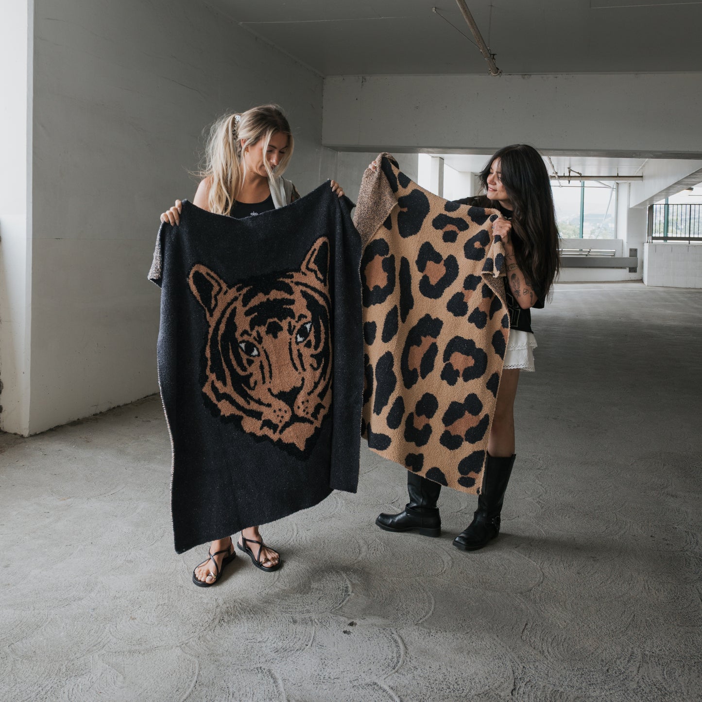 Two women stand in a parking garage, each holding up a forever french baby Plush Blanket. One features the Tiger design; the other shows leopard print. Both Oeko-Tex certified blankets are soft microfiber, and the women smile as they face each other.