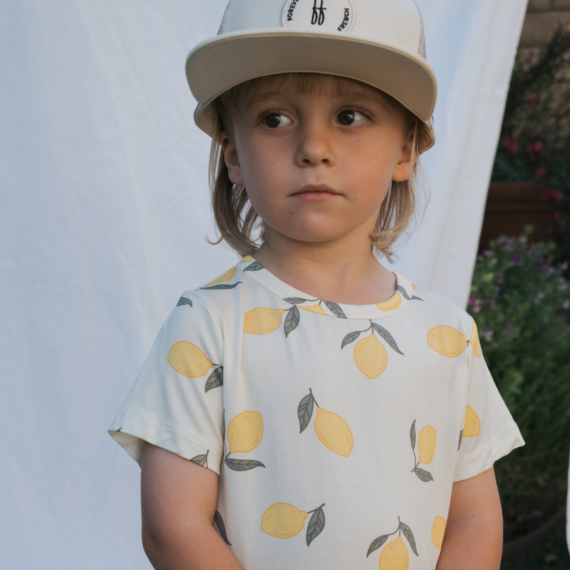 A young child models the Bamboo Short Set | Lemon by forever french baby—hypoallergenic kids clothing perfect for sensitive skin. The beige cap and white set with a yellow lemon pattern stand out against a backdrop of greenery and a white sheet.