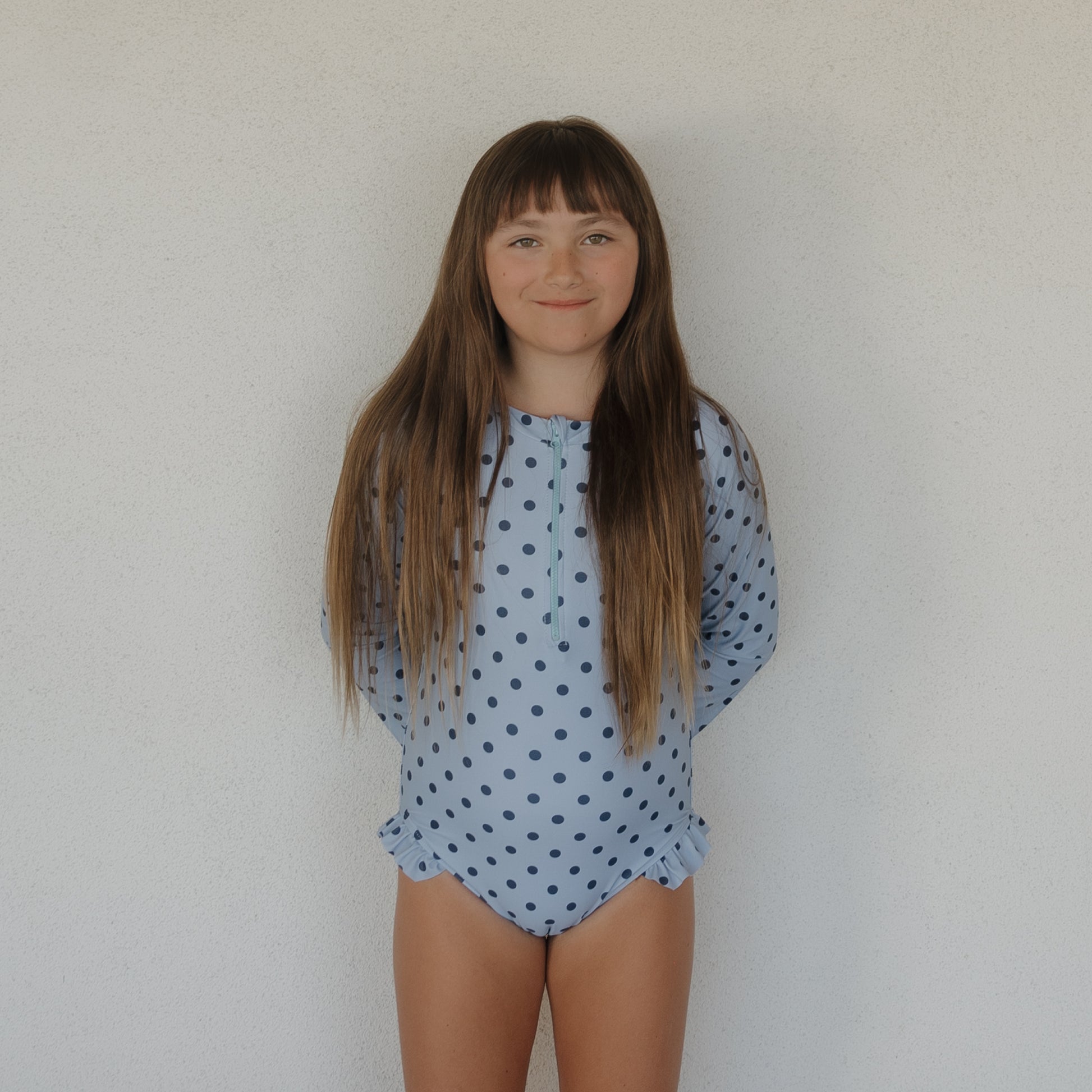 A young girl with long brown hair smiles against a white wall, wearing the forever french baby Girl's Long Sleeve Swimsuit | Sully in light blue with dark blue polka dots and ruffle details—perfect for matching family swimwear.