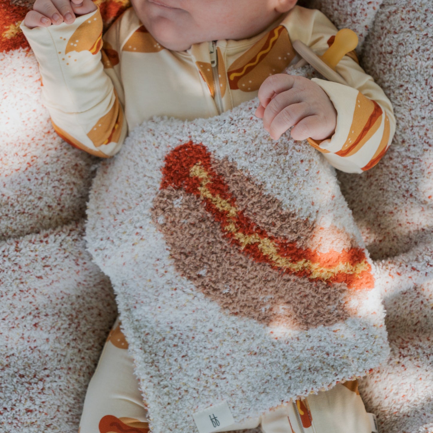 A baby in hot dog print pajamas cuddles the Lovey | Hot Dog by forever french baby, lying on soft microfiber feather yarn that matches the cozy texture of the blanket.