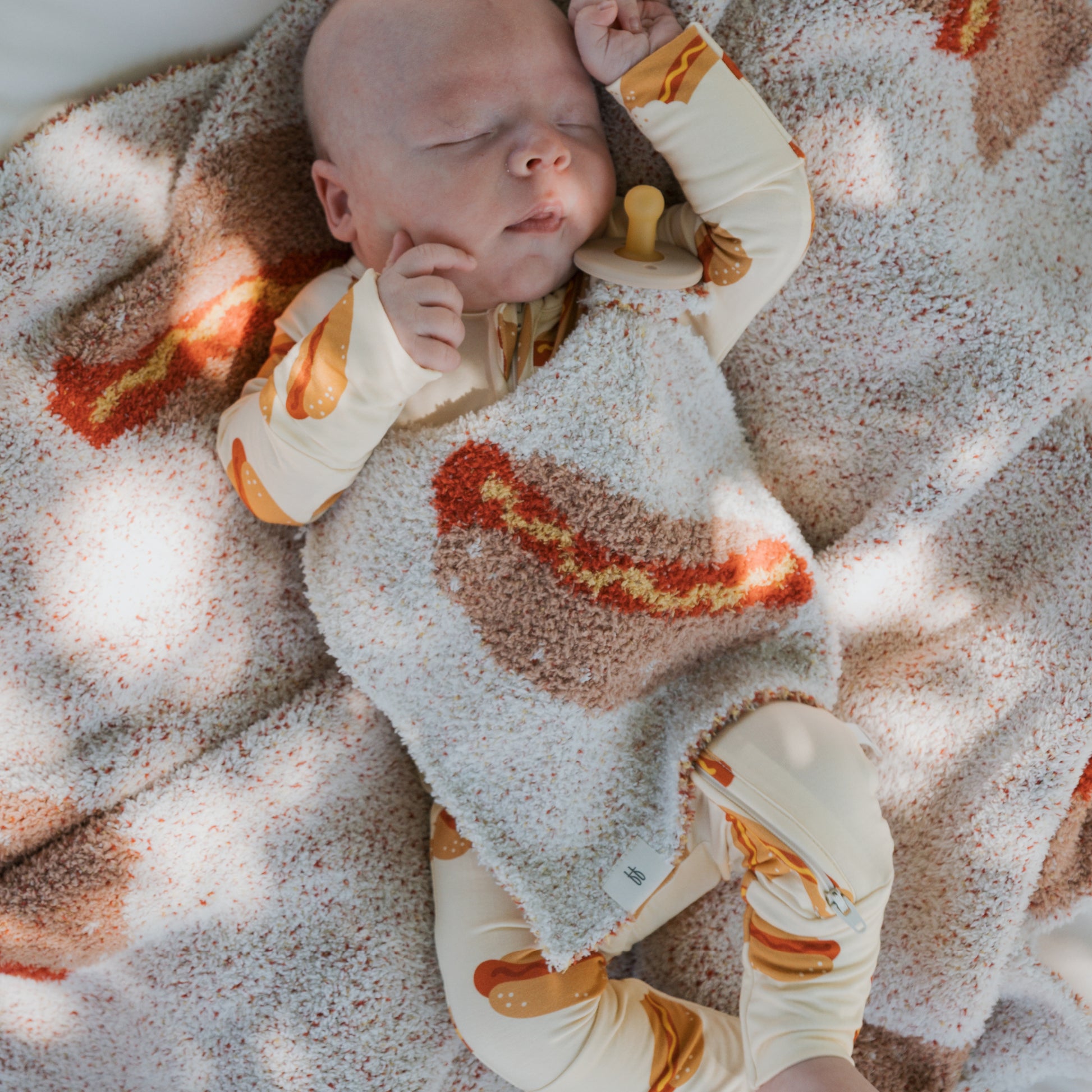 A baby in forever french baby's Bamboo Zip Pajamas | Hot Dogs sleeps soundly on a matching blanket, staying cozy and comfortable all night.