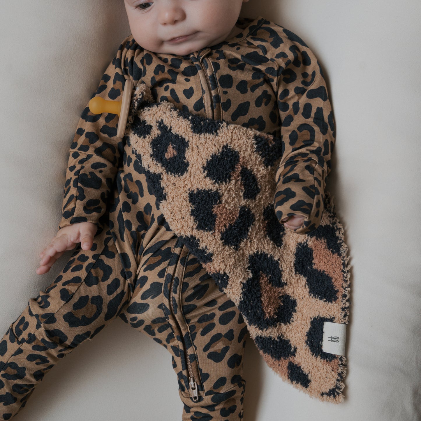 A baby wearing a brown and black leopard print outfit holds the "Lovey | Leopard" microfiber baby blanket by forever french baby while lying on a light-colored surface.