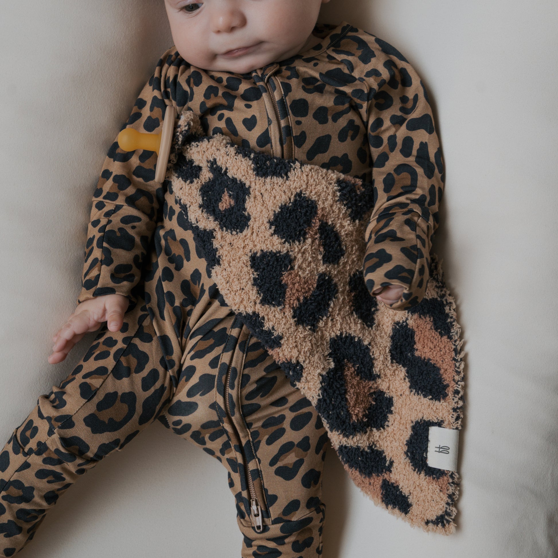 A baby wearing a brown and black leopard print outfit holds the "Lovey | Leopard" microfiber baby blanket by forever french baby while lying on a light-colored surface.