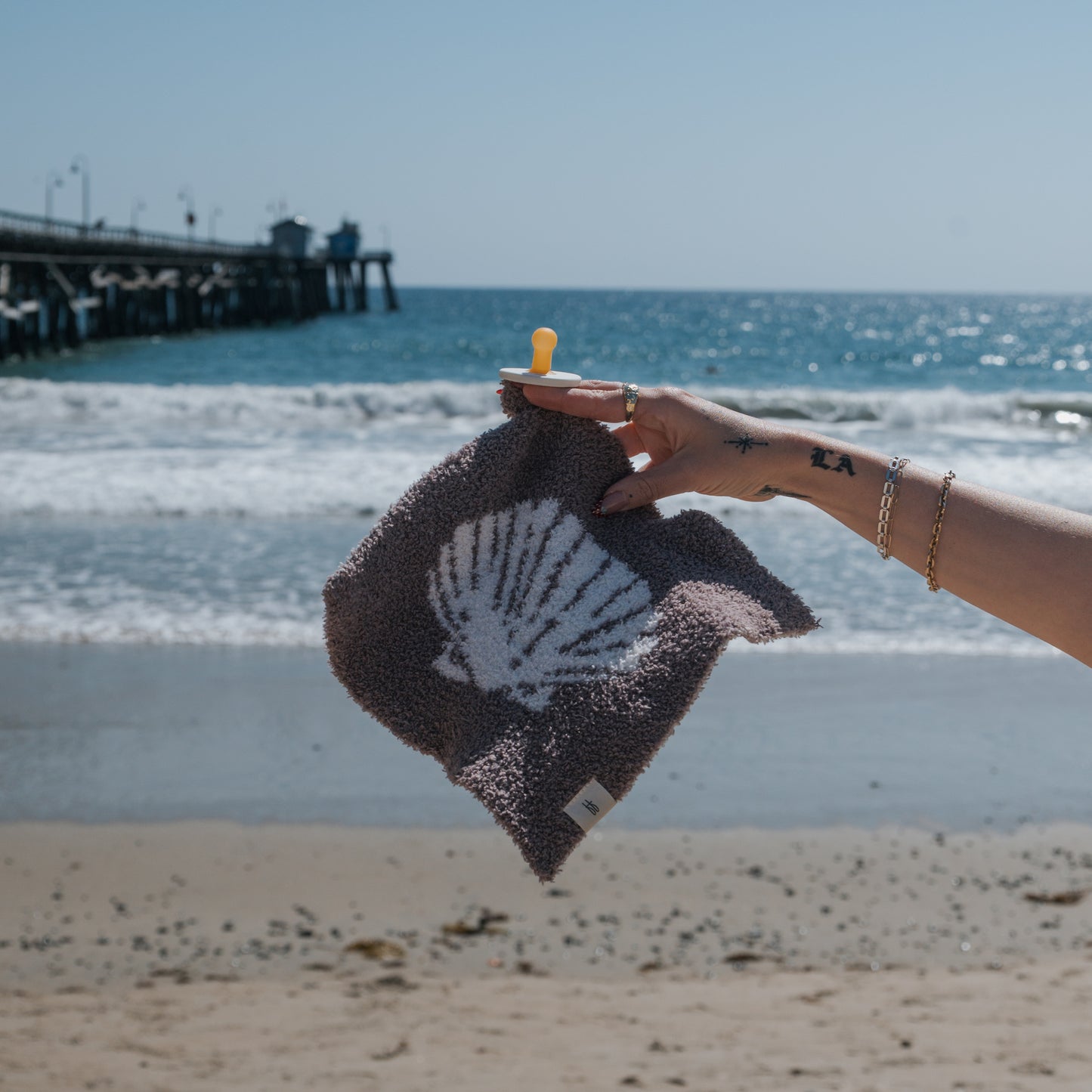 A hand holds the forever french baby Lovey | Seashells, featuring a white seashell print, with rings and bracelets gleaming on a sandy beach as waves roll in near a pier under a clear sky.