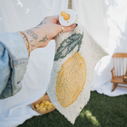 A hand holds a forever french baby *PRE-ORDER* Lovey | Lemon, made of soft microfiber feather yarn with a lemon and leaf design, against a backdrop of grass, a wooden chair, and a basket of lemons.