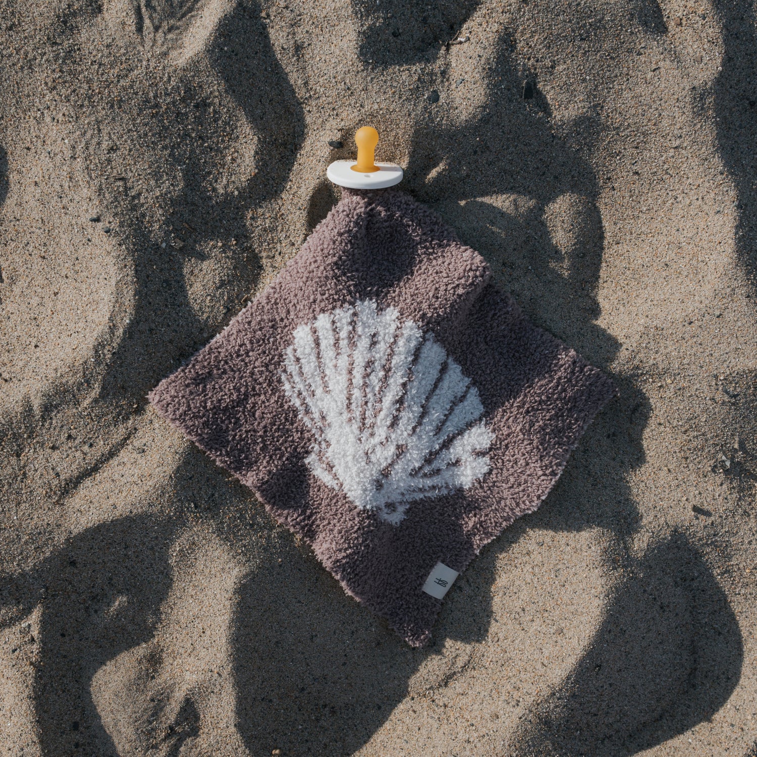 The Lovey | Seashells by forever french baby—featuring a yellow pacifier attached to a soft brown microfiber blanket with a white seashell print—rests on sand, casting gentle shadows in the sunlight.
