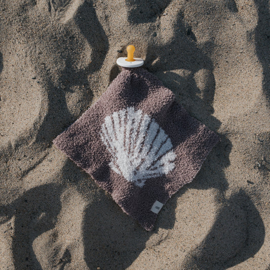 The Lovey | Seashells by forever french baby—featuring a yellow pacifier attached to a soft brown microfiber blanket with a white seashell print—rests on sand, casting gentle shadows in the sunlight.