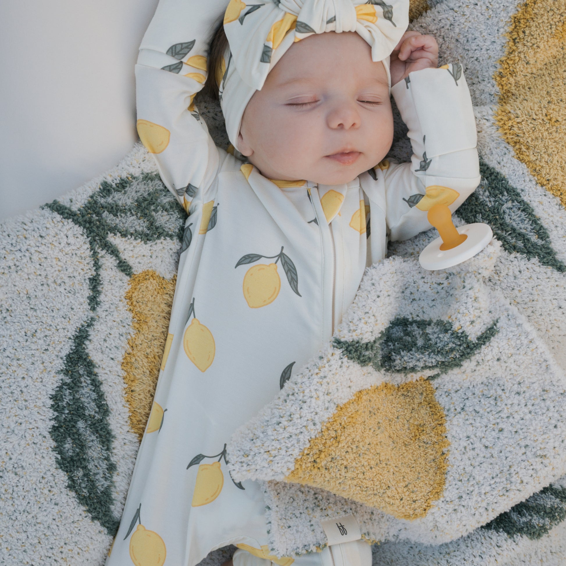 A baby sleeps on a blanket with yellow lemons, wearing a lemon-patterned forever french baby onesie and headband. Nearby are a pacifier and the *PRE-ORDER* Lovey | Lemon from forever french baby as the baby rests with arms raised.