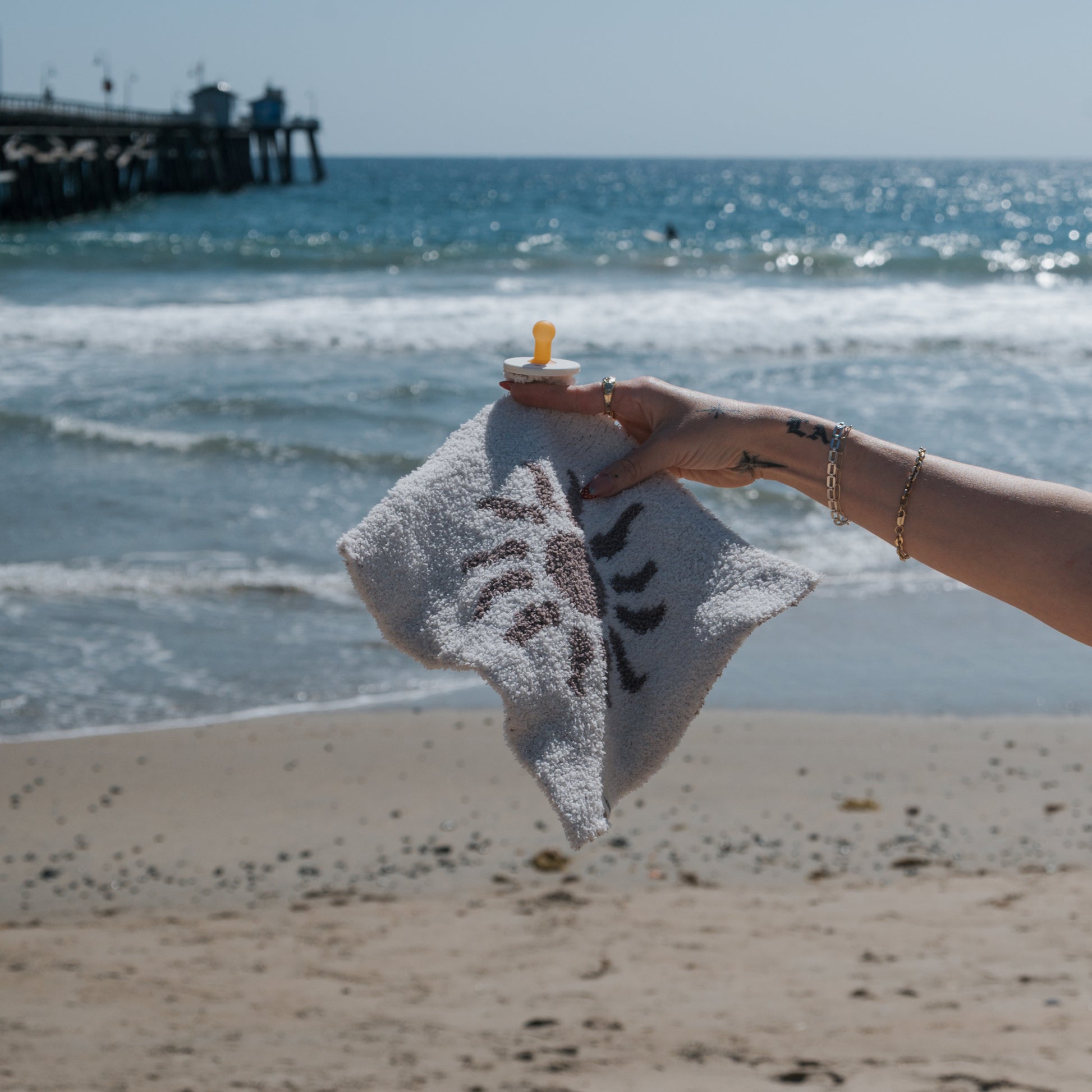 A hand holds the forever french baby Lovey | Summer Sun—a soft microfiber blanket with a cheerful sun print and attached yellow pacifier—against an ocean backdrop with sandy beach and pier.