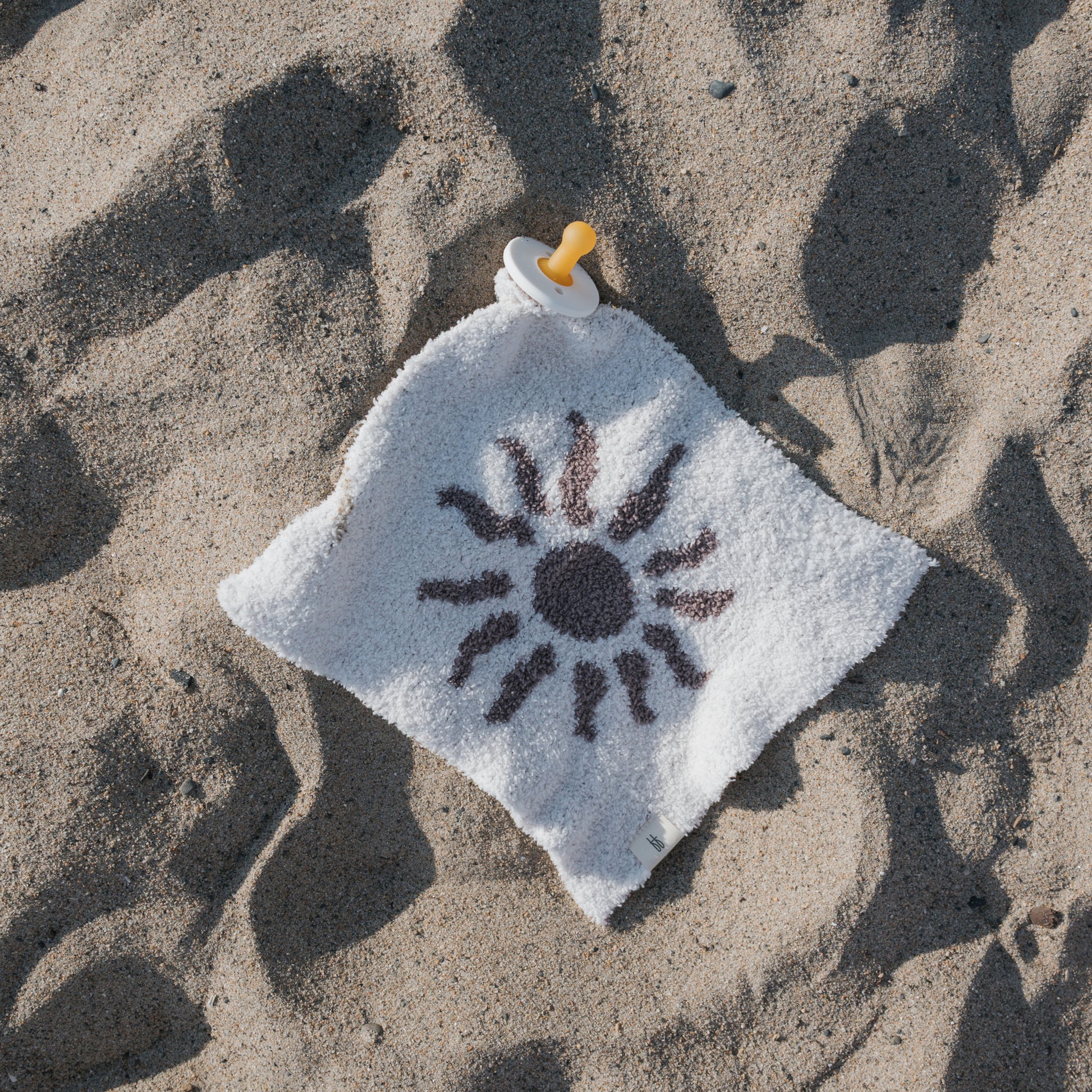 The forever french baby Lovey | Summer Sun, a white square microfiber baby blanket with an attached pacifier and sunny print, rests on sandy ground in the sunlight—perfect for warm days.