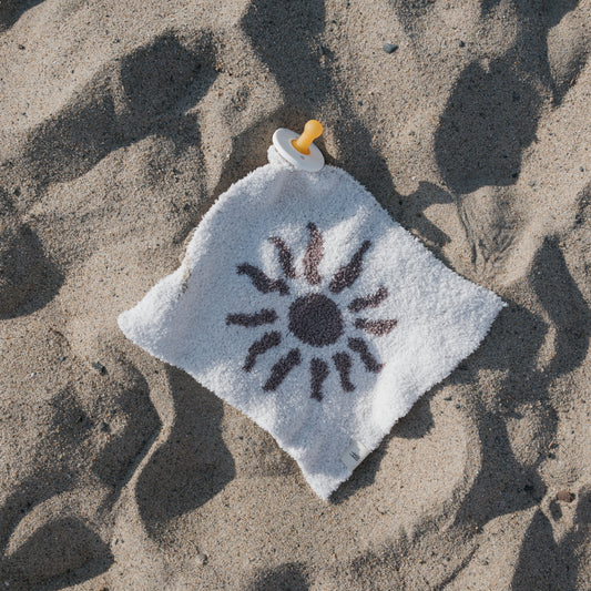The forever french baby Lovey | Summer Sun, a white square microfiber baby blanket with an attached pacifier and sunny print, rests on sandy ground in the sunlight—perfect for warm days.