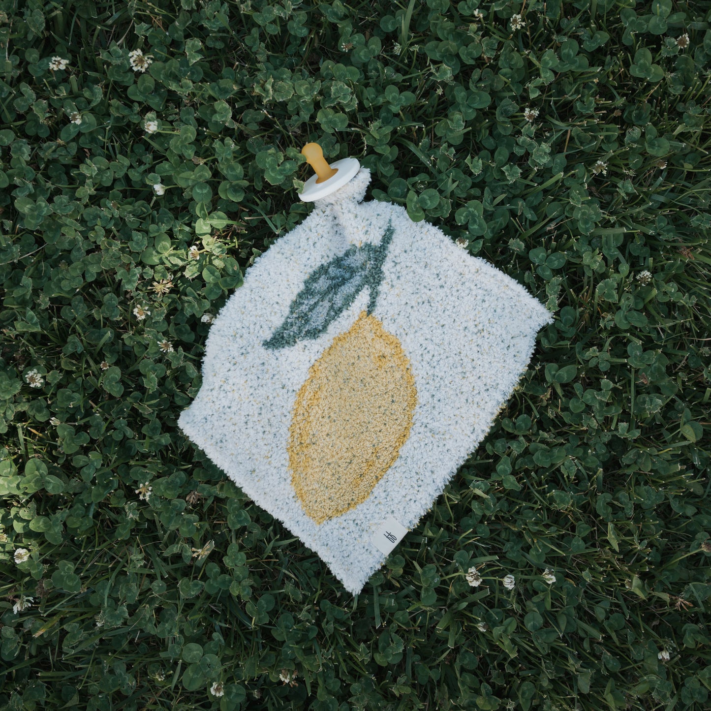 The *PRE-ORDER* Lovey | Lemon by forever french baby features a soft microfiber feather yarn lovey with an embroidered yellow lemon and green leaf, attached to a baby pacifier and displayed on green grass with white clover flowers.