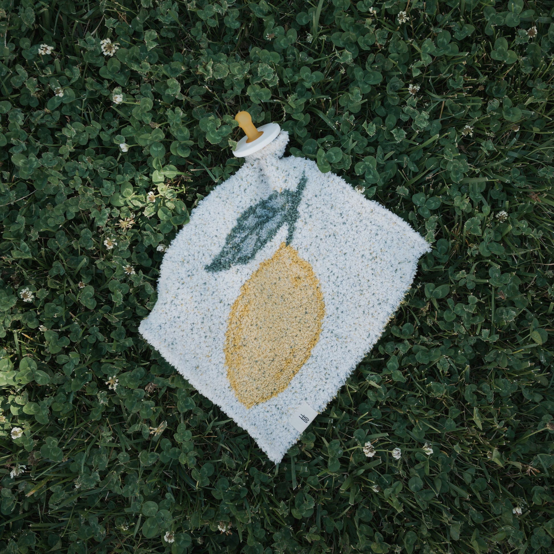 The *PRE-ORDER* Lovey | Lemon by forever french baby features a soft microfiber feather yarn lovey with an embroidered yellow lemon and green leaf, attached to a baby pacifier and displayed on green grass with white clover flowers.