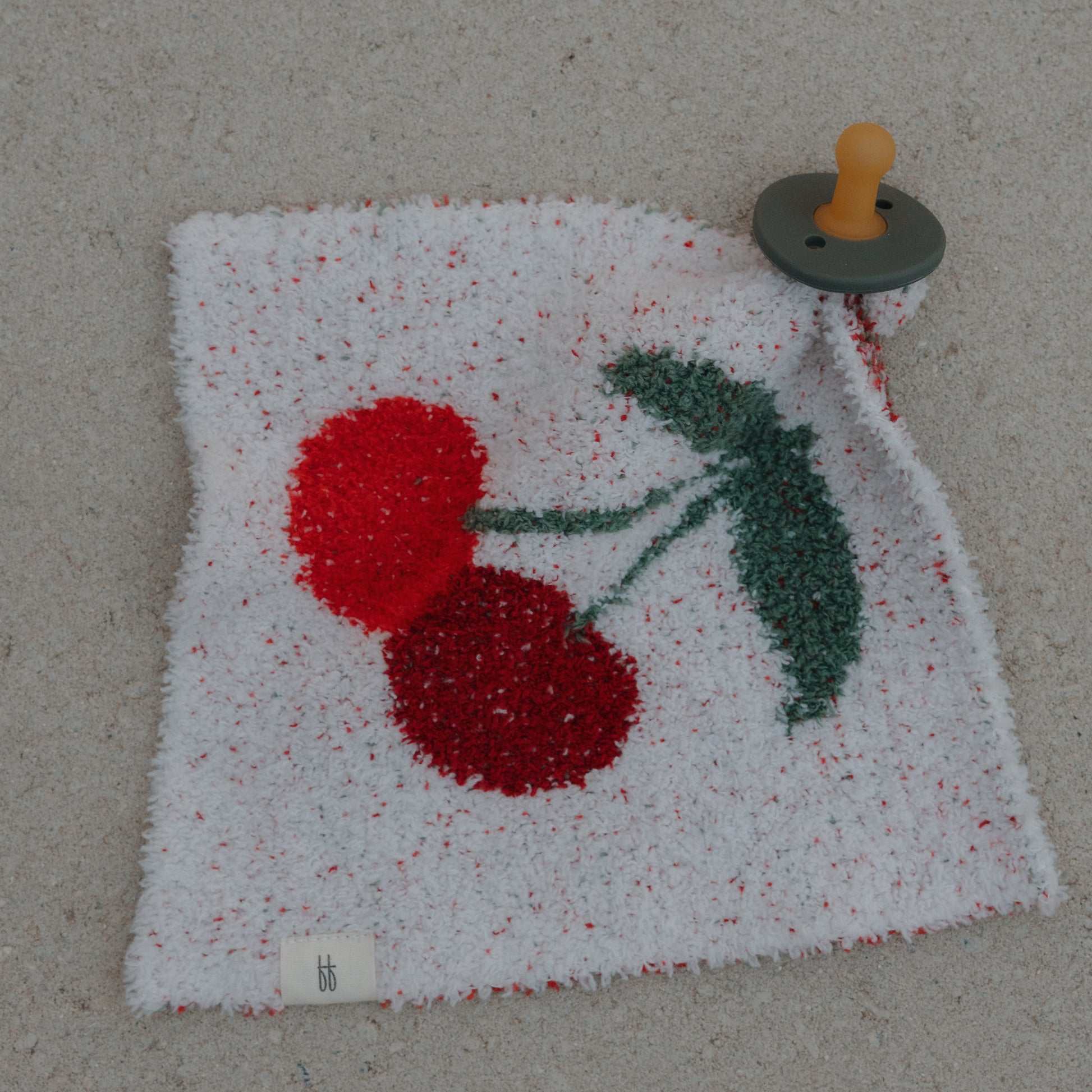 The forever french baby Lovey | Cherry is a soft, white microfiber security blanket with a cherry print and an attached pacifier, featuring a gray base and tan nipple, shown lying on a light surface.