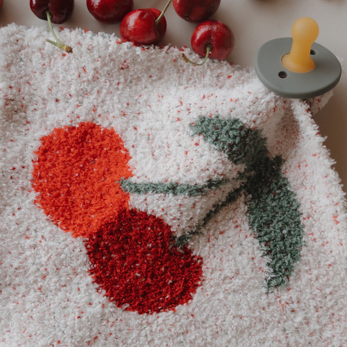 A close-up of the forever french baby Lovey | Cherry, a soft white microfiber blanket with cherry print, displayed next to real cherries and a gray pacifier with yellow nipple on a light surface.