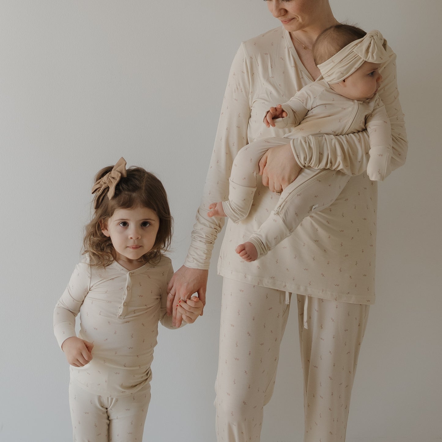 A woman wears forever french baby Bamboo Two Piece Pajamas | Lucy in cream, holding a baby and the hand of a young girl, both also in matching bamboo sleepwear, all standing together against a plain light background.