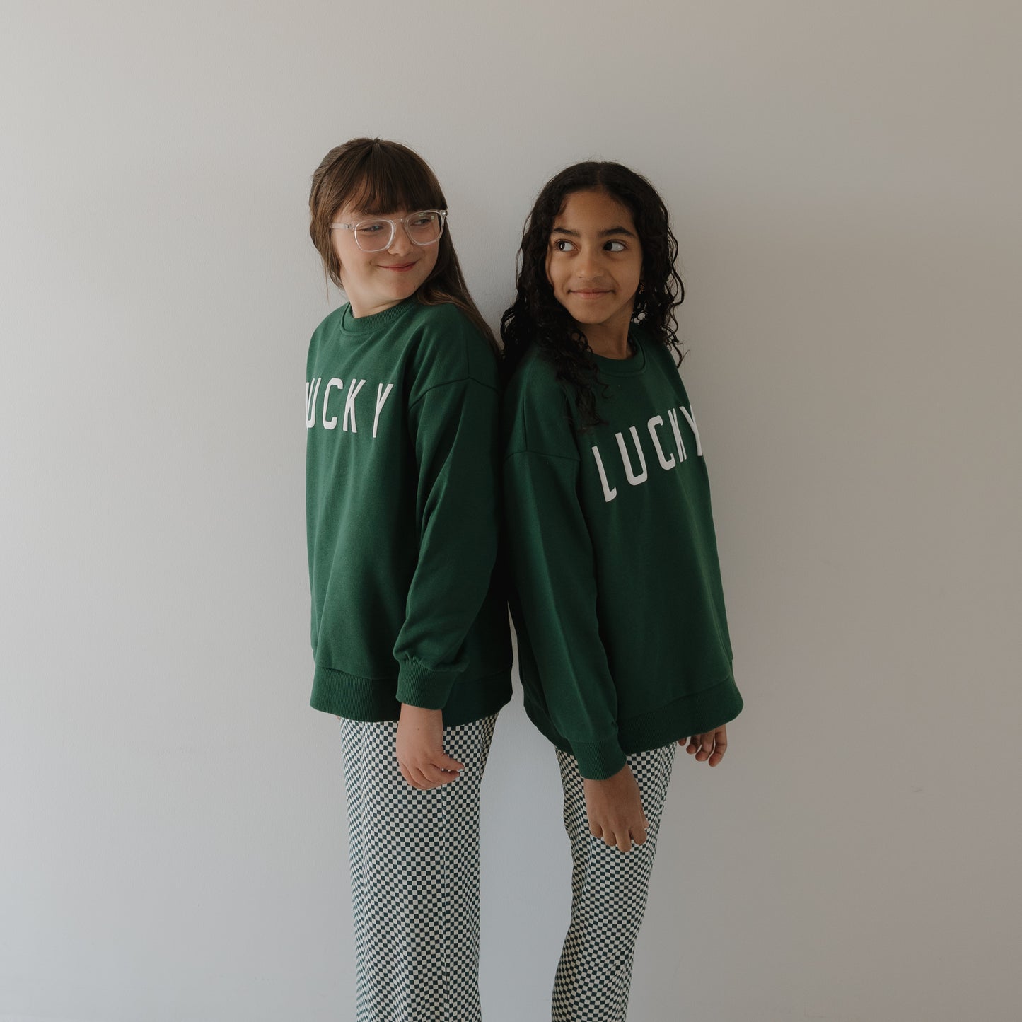 Two girls smile back-to-back in forever french baby’s Child Sweatshirt | Lucky and black-and-white checkered pants—cozy matching outfits for St. Patrick's Day, set against a simple light background.
